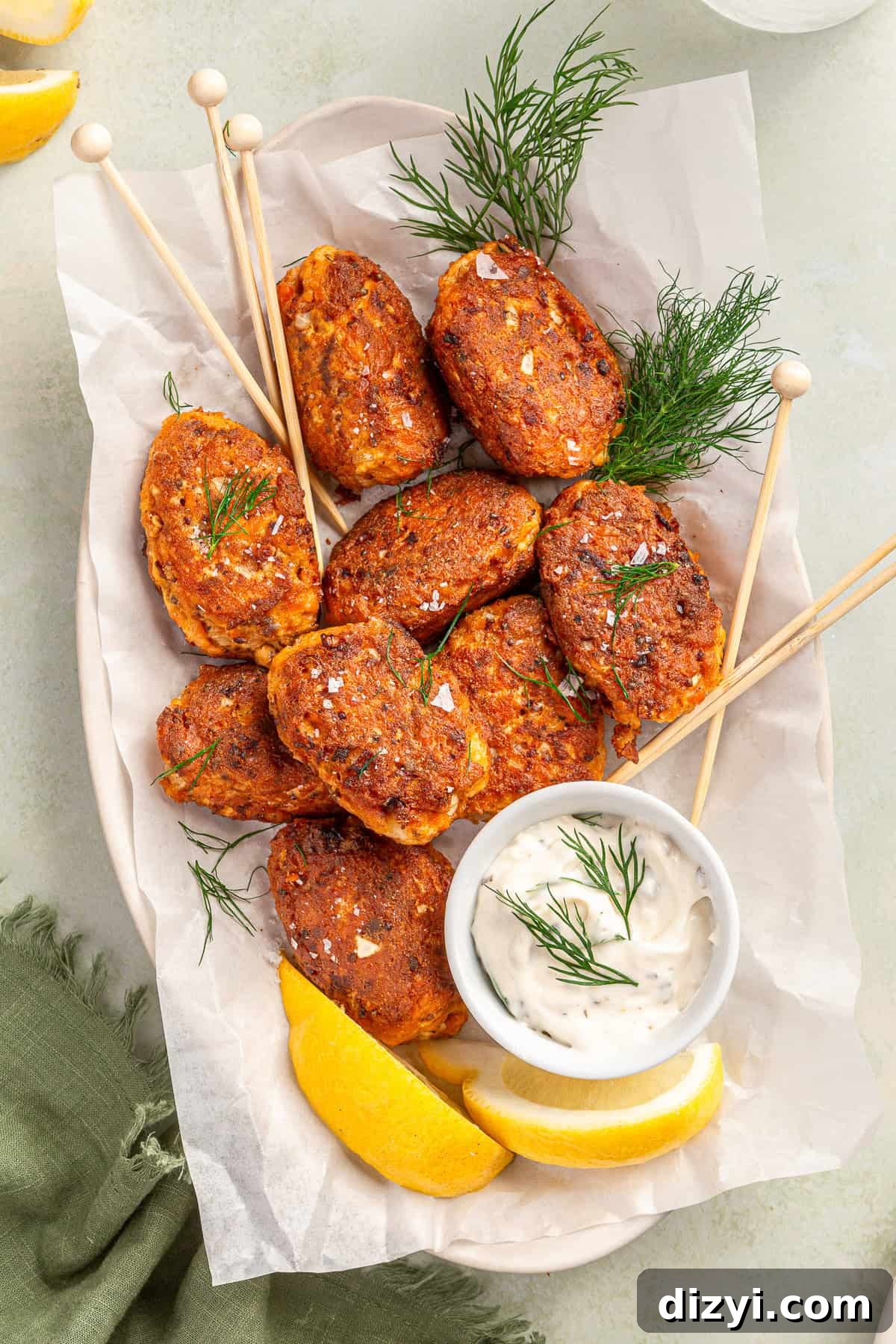 A basket of finished salmon croquettes on a piece of white parchment paper. Skewers, a small bowl of sauce, lemon wedges, and dill sprigs are also in the image.