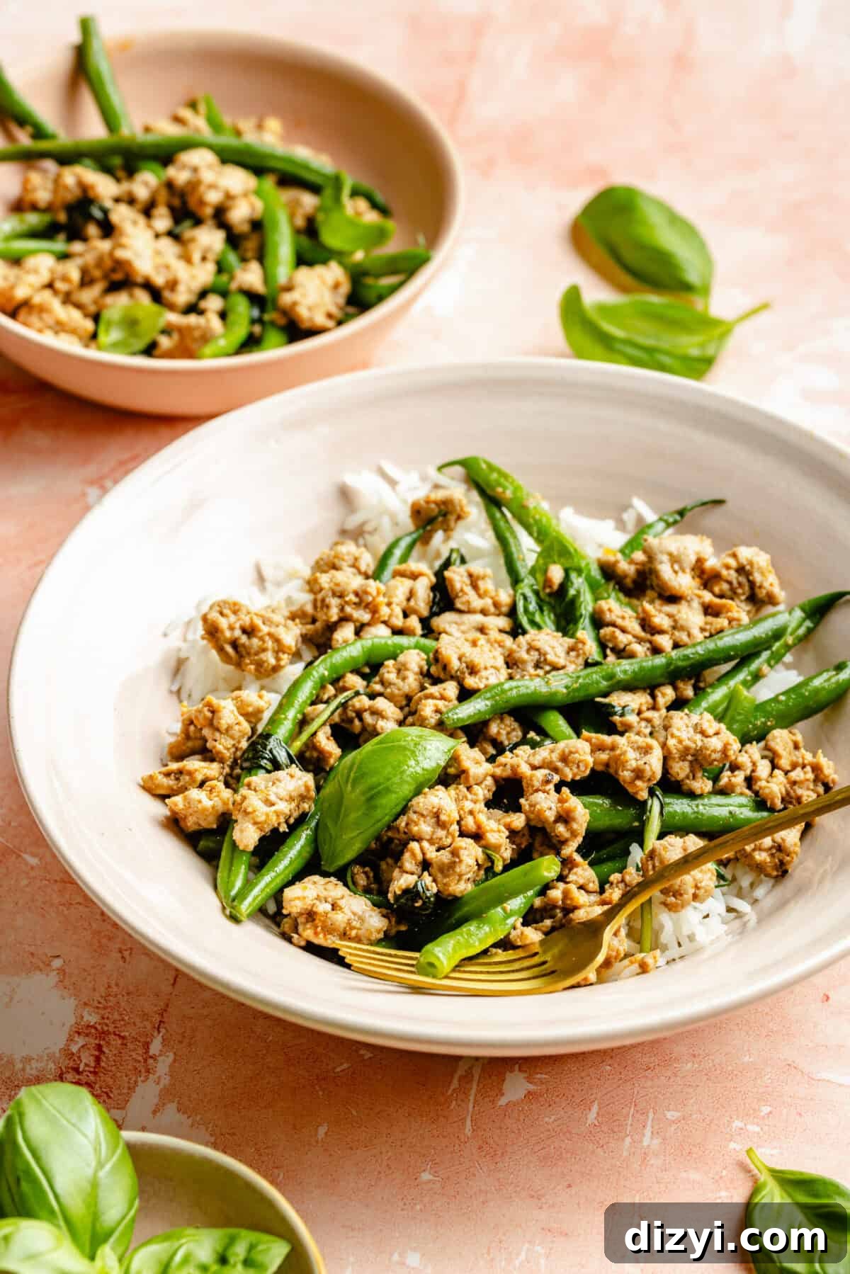 The turkey, green bean, and basil dish served on a bed of white rice, in a white bowl with basil leaves on the table and a second dish behind.