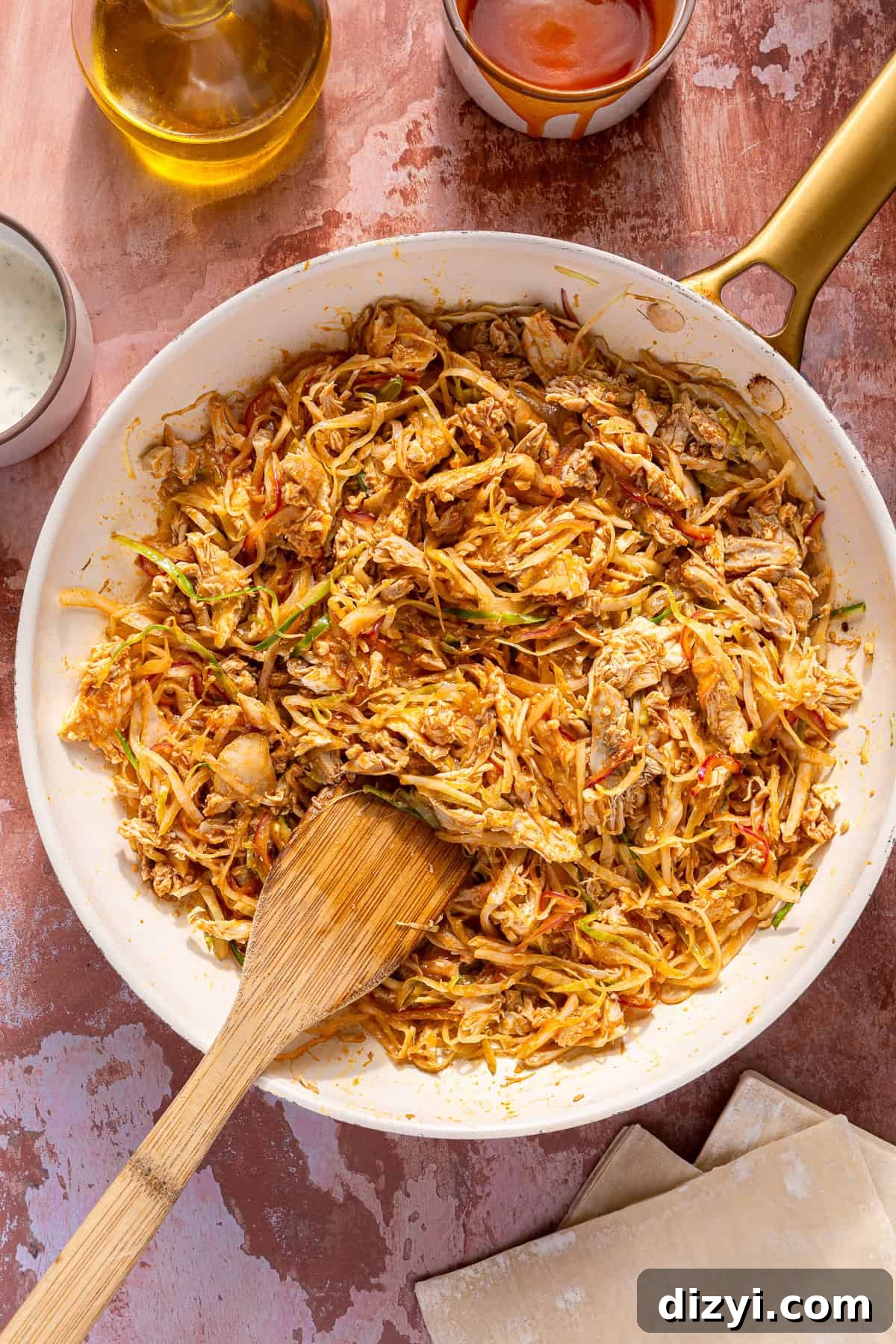 The onion, cabbage, chicken, and sauce needed for Buffalo Chicken Egg Rolls, being cooked in a white pan with a wooden spoon.