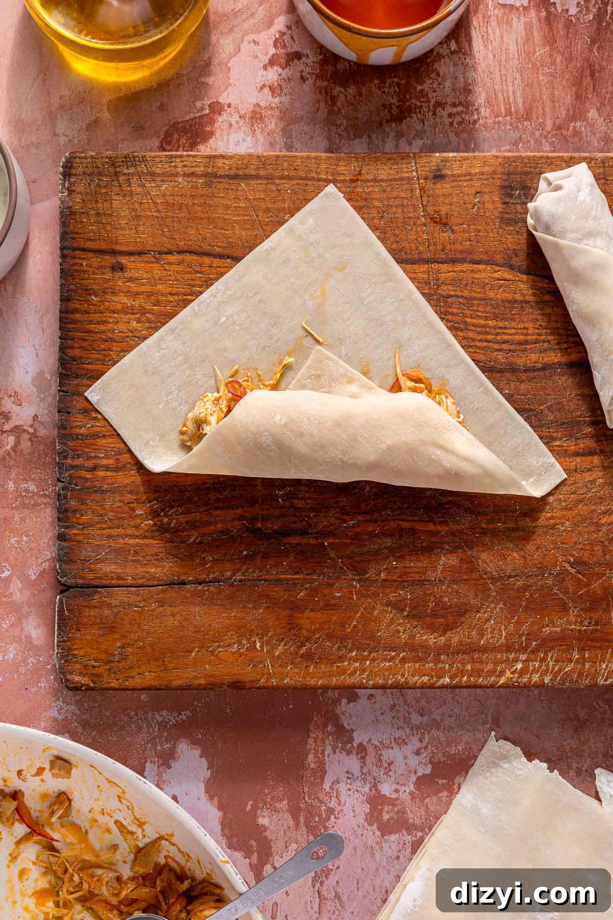 An egg roll wrapper, laid on a wooden cutting board, with the Buffalo Chicken Egg Roll mixture inside. It is in the process of being rolled.