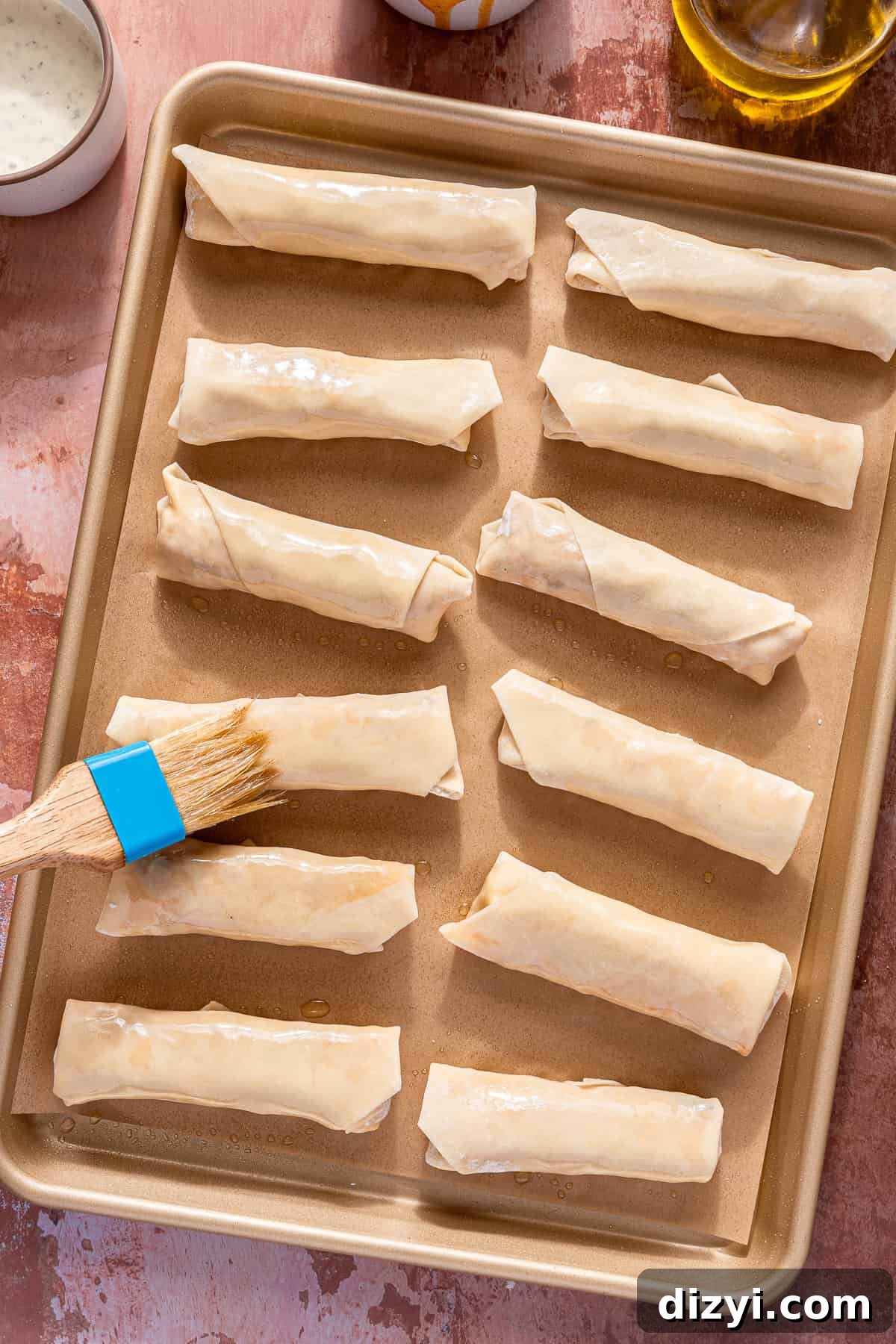 A sheet pan of twelve Buffalo Chicken Egg Rolls being brushed with avocado oil.