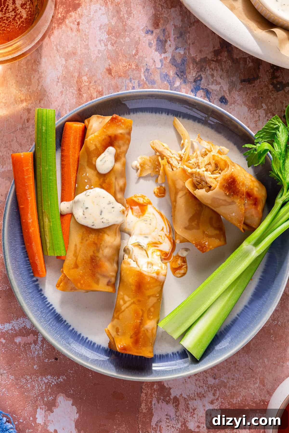A white and blue plate containing three finished Buffalo Chicken Egg Rolls, a few sticks of celery and carrots, and some ranch drizzled on top.