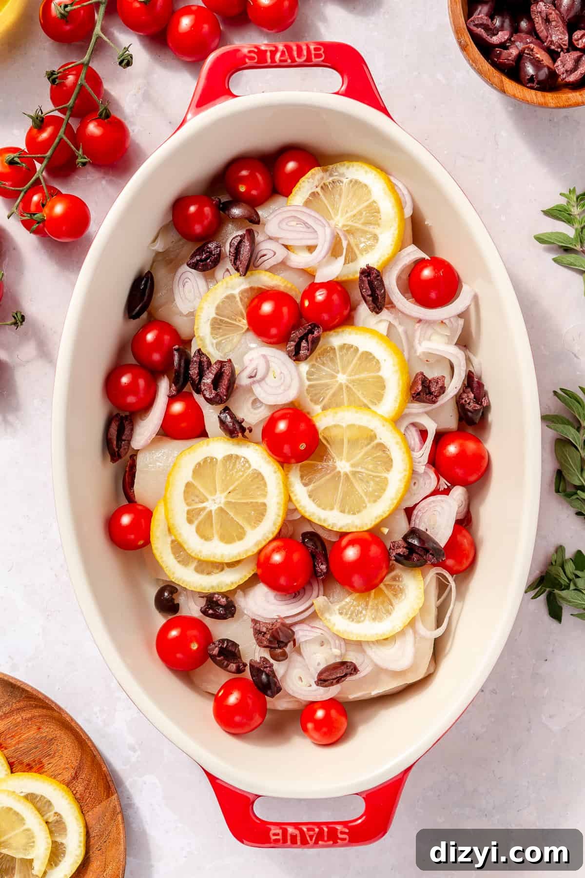 Raw halibut in oval baking dish with shallot, lemon, olives, and onions scattered on top.