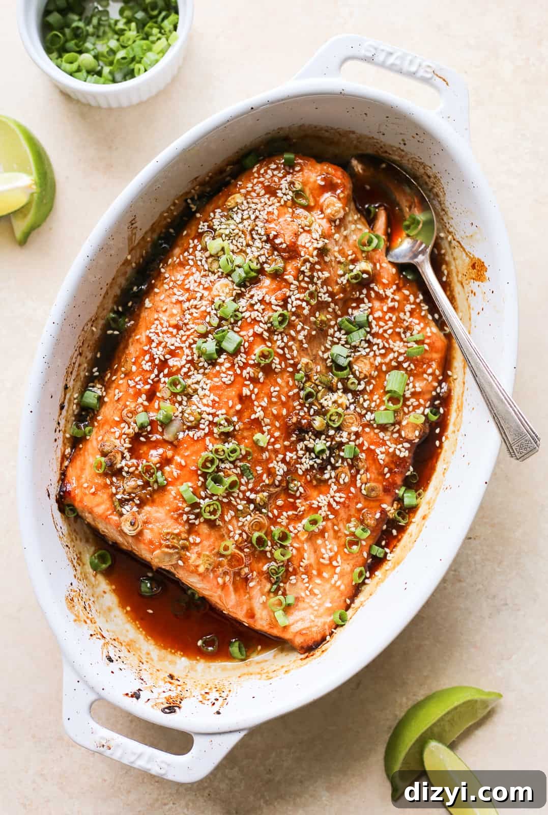Large salmon filet in white oval baking dish covered in marinade, sesame seeds, and sliced green onions.