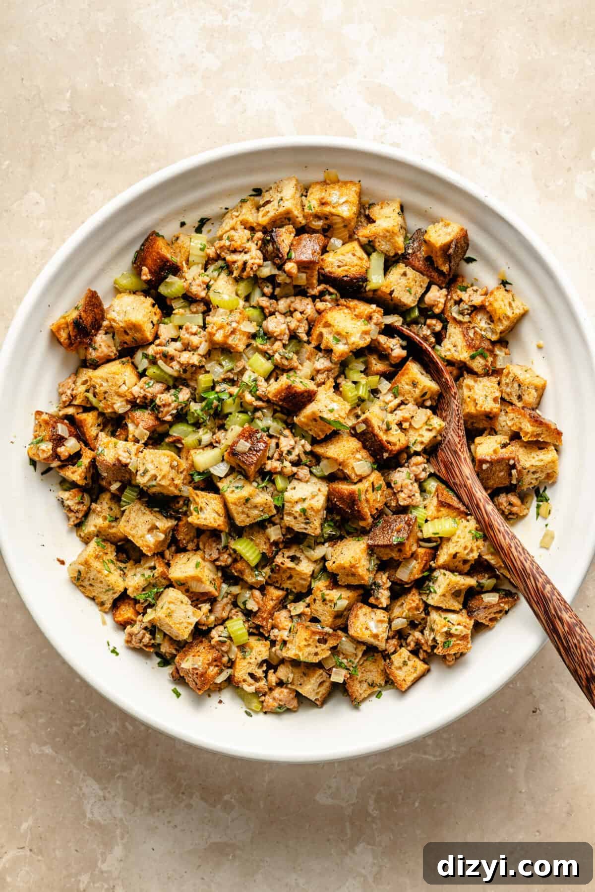 Oven-Baked Sausage and Sage Stuffing 5 a large white mixing bowl with bread cubes, herbs, celery, and sausage on a neutral background