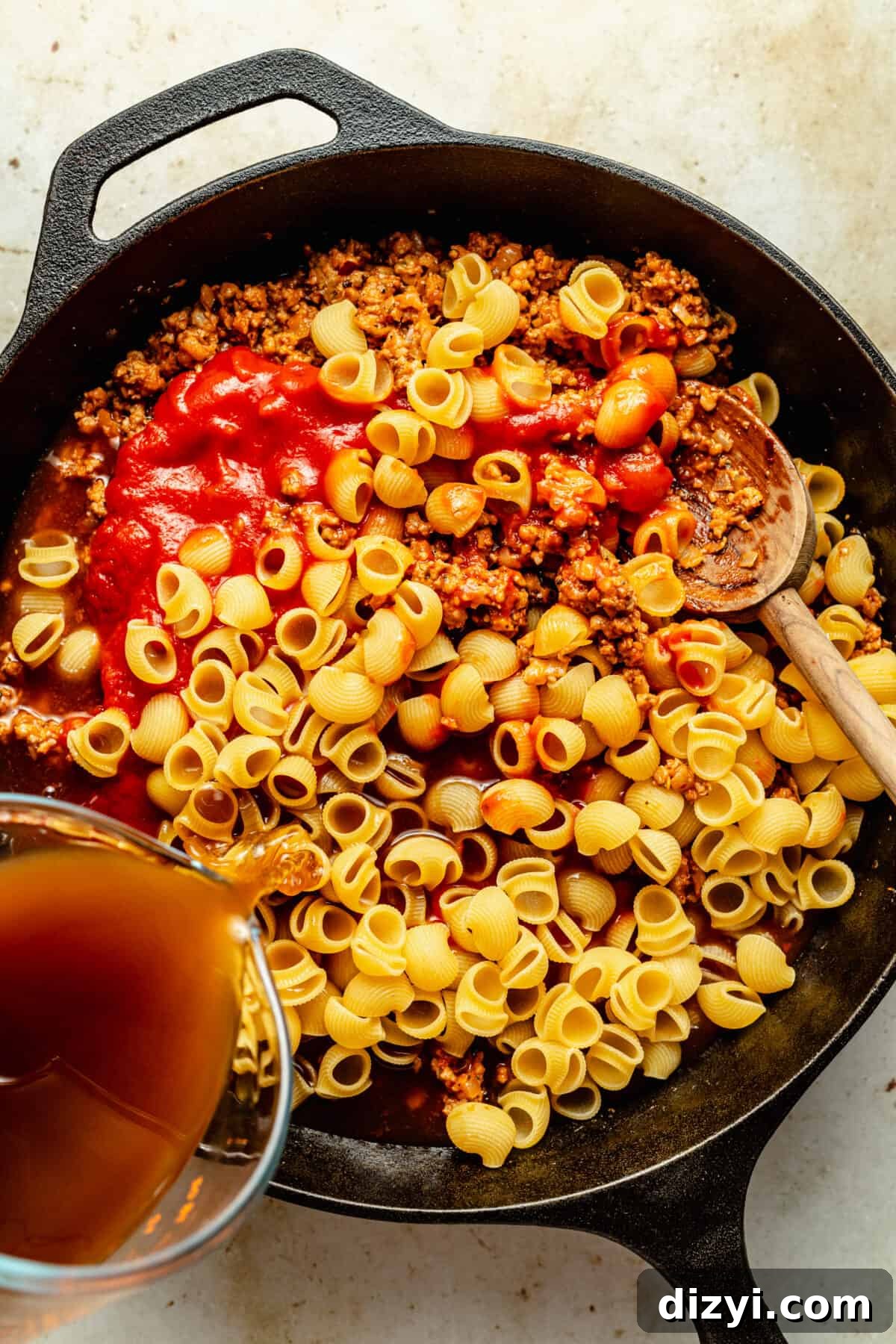 Black cast iron skillet filled with browned ground beef, dry pasta shells, red sauce and broth pouring in.