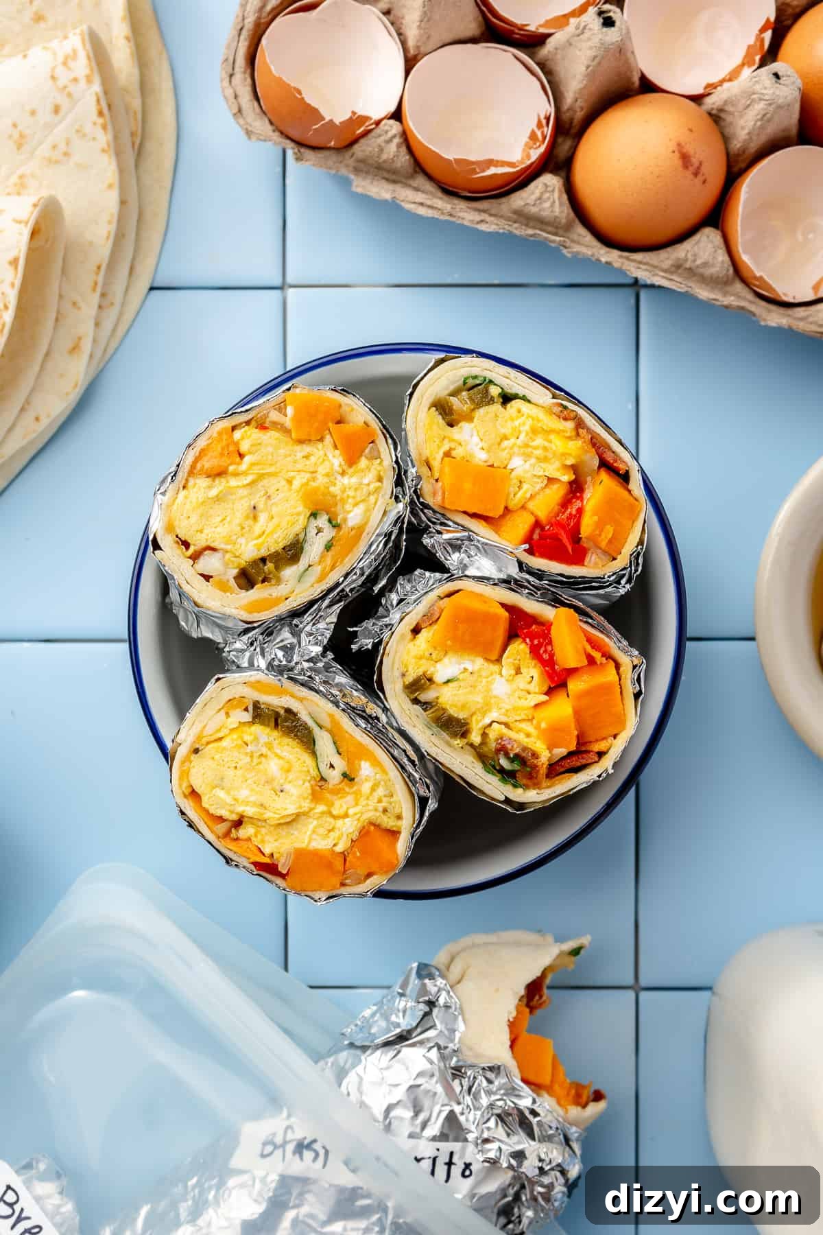 Burritos cut in half and facing up on a plate in the center of photo with cracked eggs in corner and another burrito at the bottom. All on blue tile.