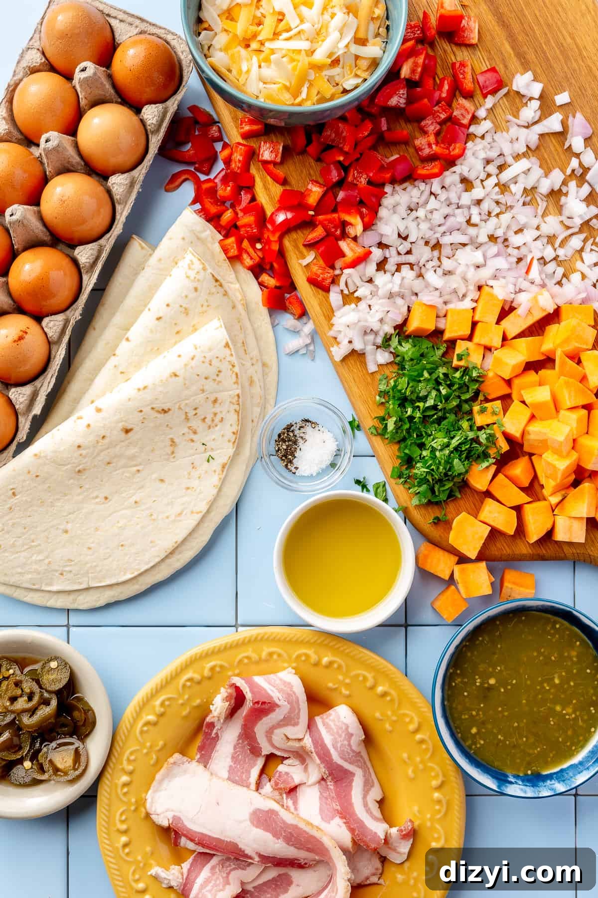Breakfast burrito ingredients. Whole eggs, tortillas, jalapeños, shredded cheese, diced red bell pepper, diced shallot, diced sweet potatoes, chopped herbs, bowl of salsa verde, and plate with uncooked bacon strips on blue tile.