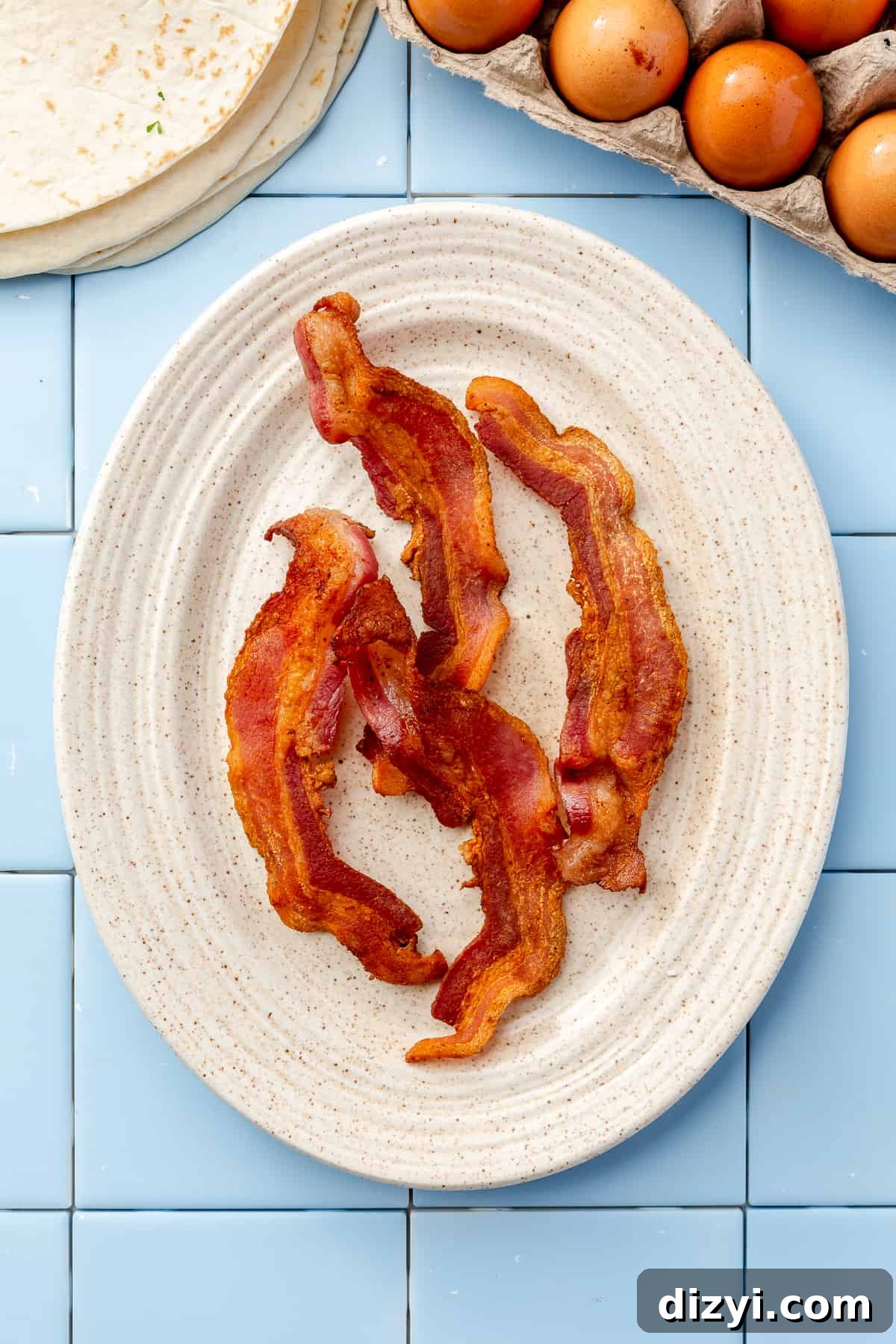 Oval cream plate filled with 4 pieces of crisped bacon on blue tile backdrop.