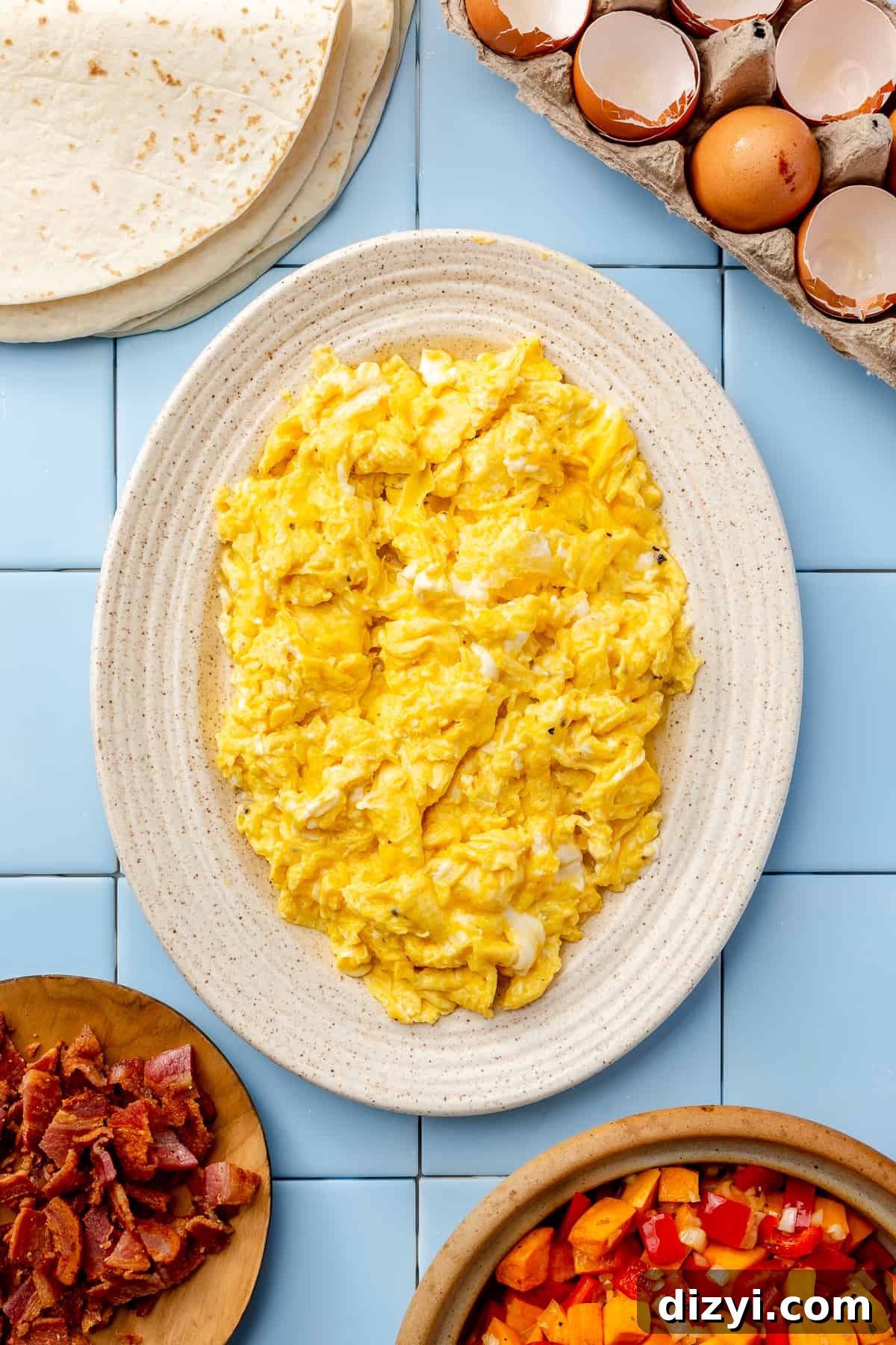 Oval cream plate filled with scrambled eggs. Cracked eggs in top corner and bacon in other corner.