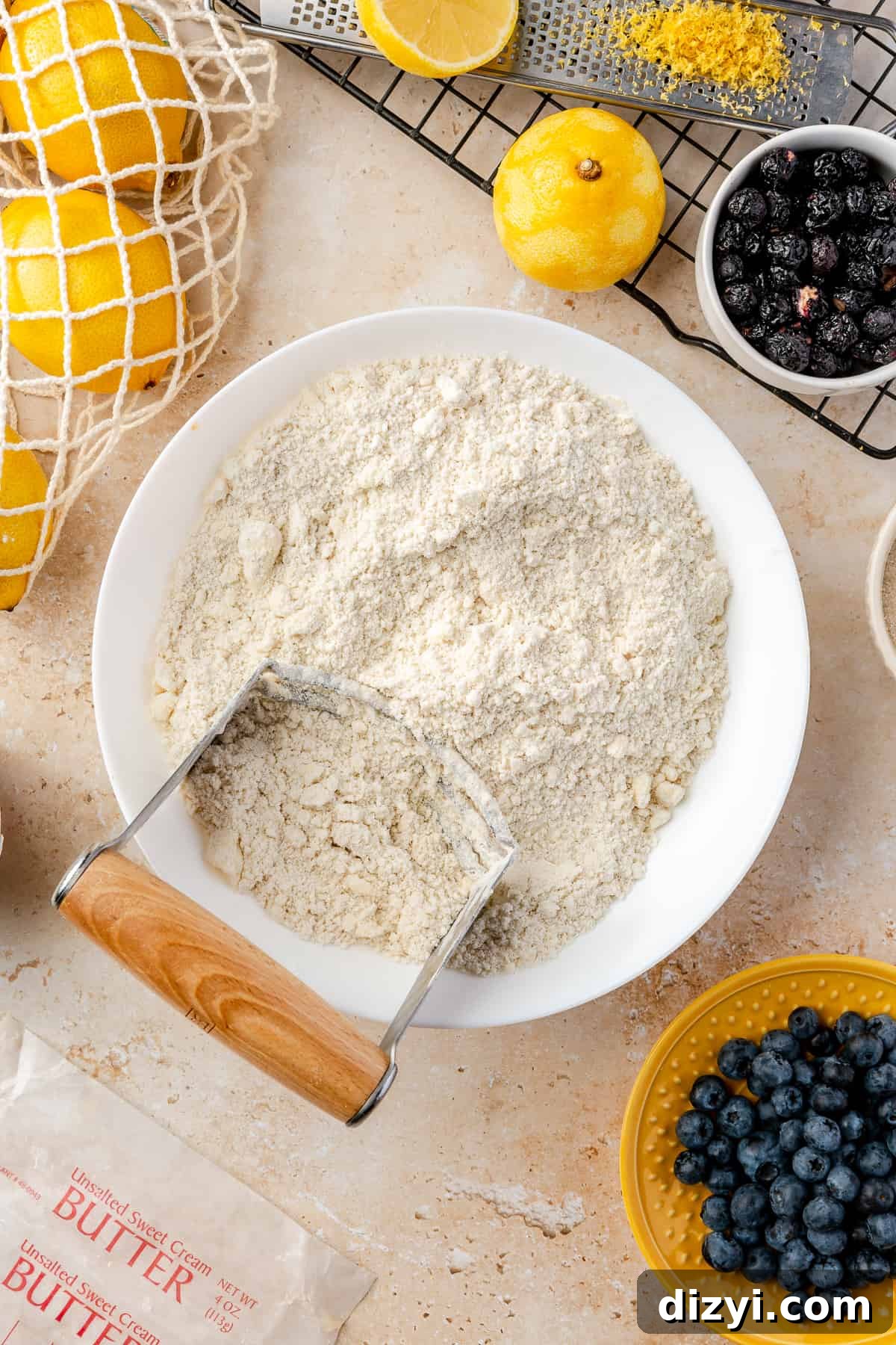 Flour and cut butter in large white bowl with other scone ingredients scattered around it. Pastry cutter resting in bowl.