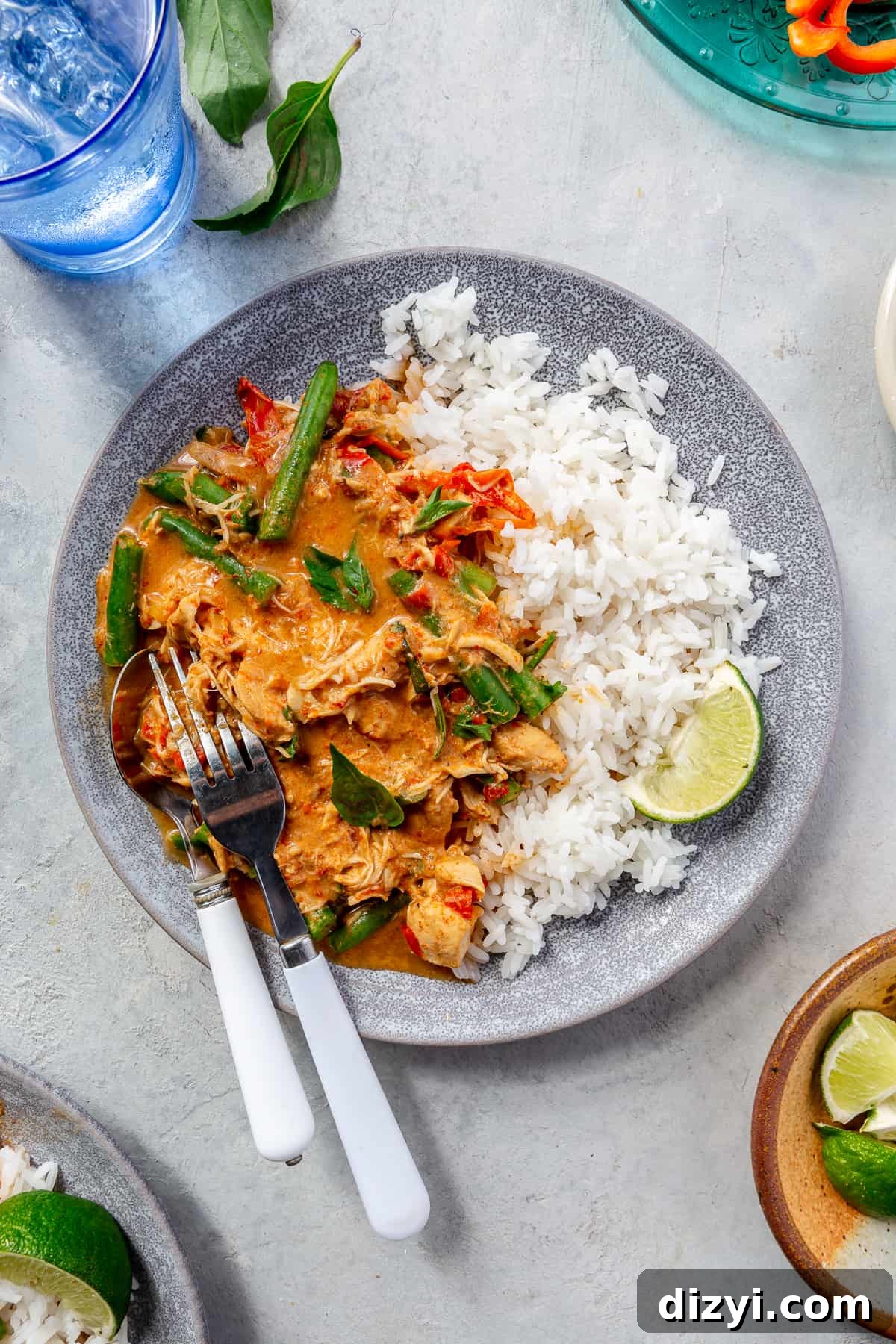 Slow Cooker Thai Red Curry with Chicken and Vegetables 2 Blue bowl filled with Slow Cooker Thai Red Curry Chicken and Veggies and rice. Cup of water and bowls of garnishes surrounding.