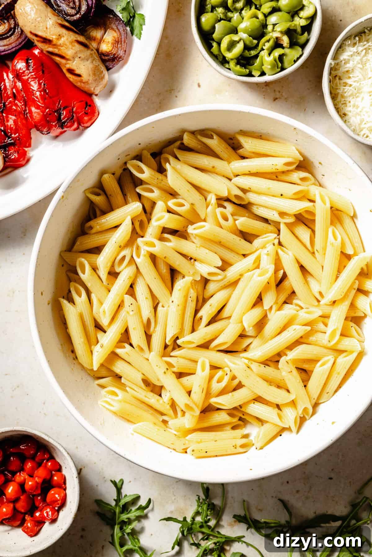 Cooked pasta in large bowl with vinaigrette. Additional Grilled Antipasto Pasta Salad ingredients scattered around.