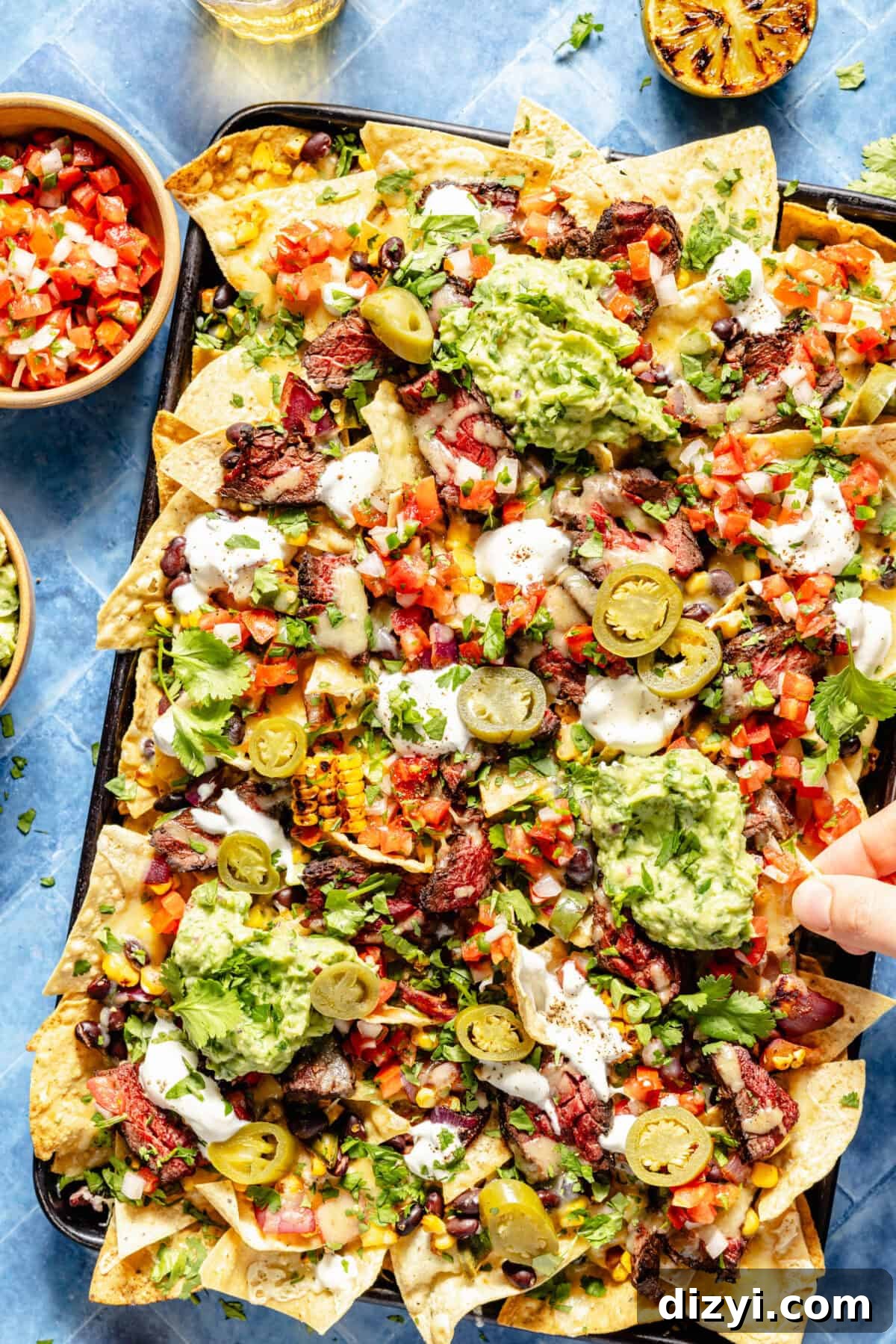 Grilled Chipotle Steak Nachos assembled on sheet pan. Hand grabbing a chip.