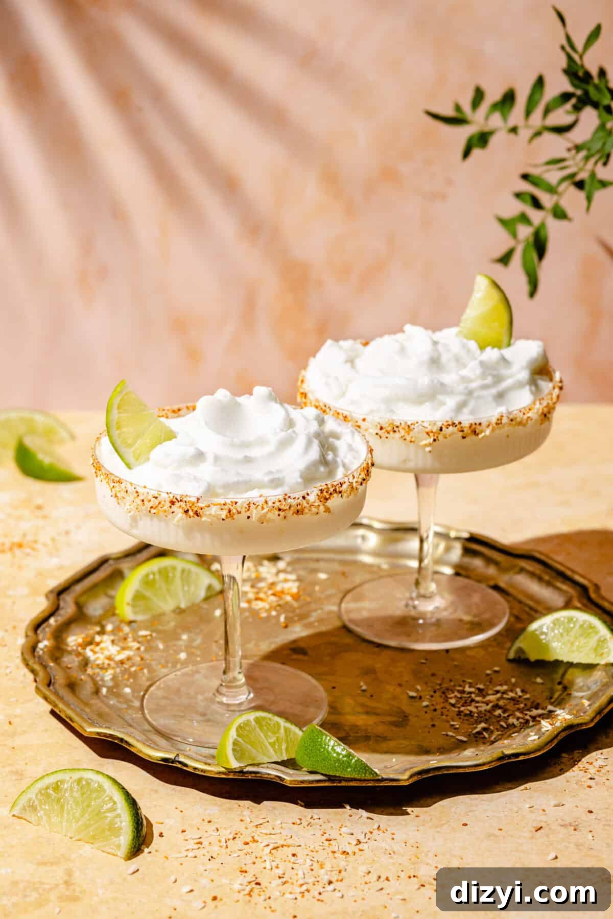 Two Frozen Coconut Margaritas in coupe cocktail glasses, each garnished with a lime wedge. The margaritas sit on a bronze tray on top of a tan table, with lime wedges on both the tray and the table. A green branch is visible in the top right corrner.
