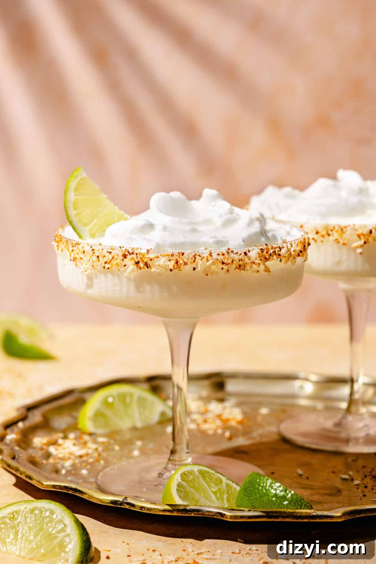 Straight shot of two coupe cocktail glasses, each containing a frozen coconut margarita. The glass in the forefront is garnished with a lime wedge, and there are limes on both the bronze tray the glasses are on, as well as on the table.