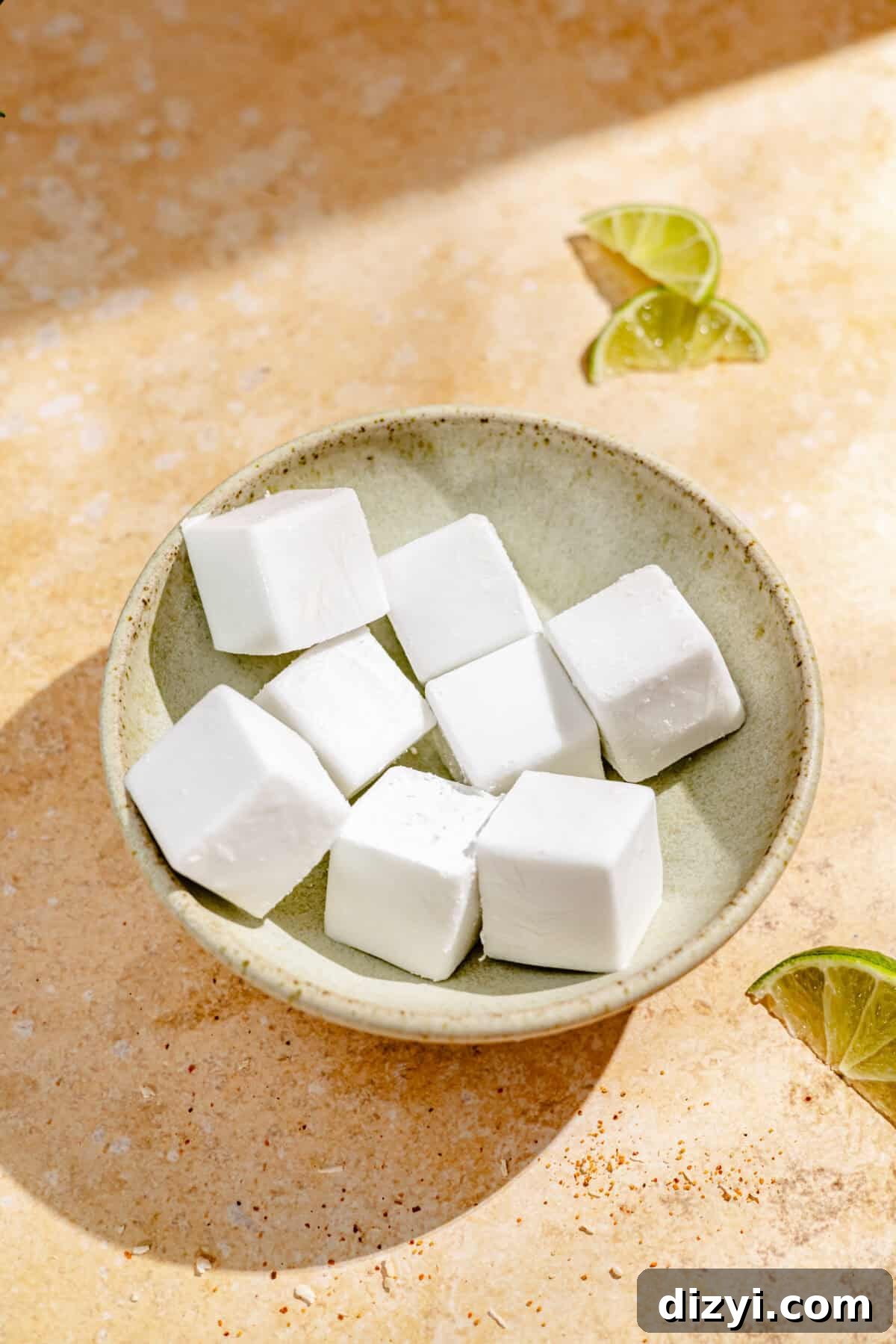 A beige bowl with cubes of frozen coconut milk. Three lime wedges are visible, and the bowl rests on a tan table.
