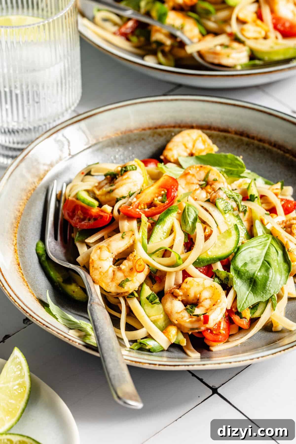 Two cream bowls with assembled Tangy Thai Shrimp Noodle Salad. Ribbed glass of ice water in the corner. Lime wedges in the bottom left corner.