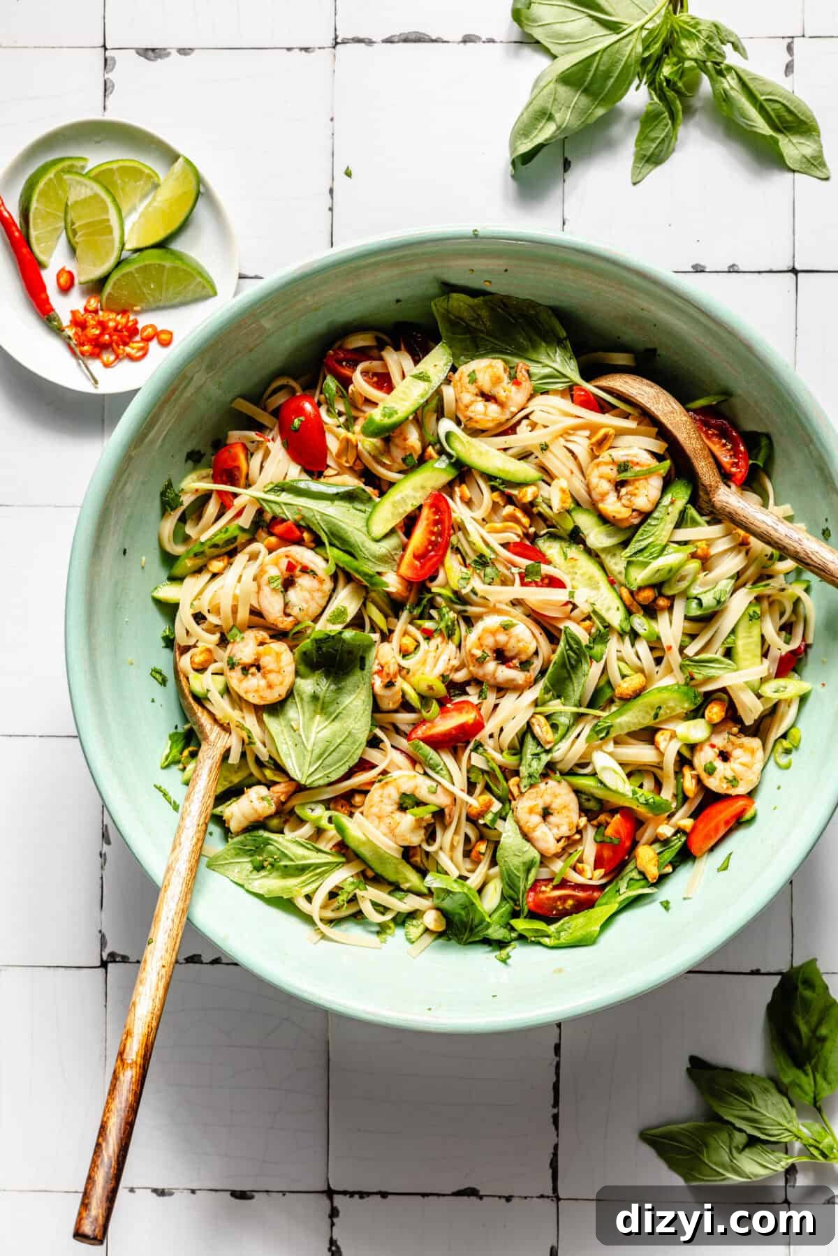 Sage green serving bowl of Tangy Thai Shrimp Noodle Salad, with wooden tongs. Basil leaves on the top and bottom of the image, and a bowl of limes and Thai chiles in the top left.