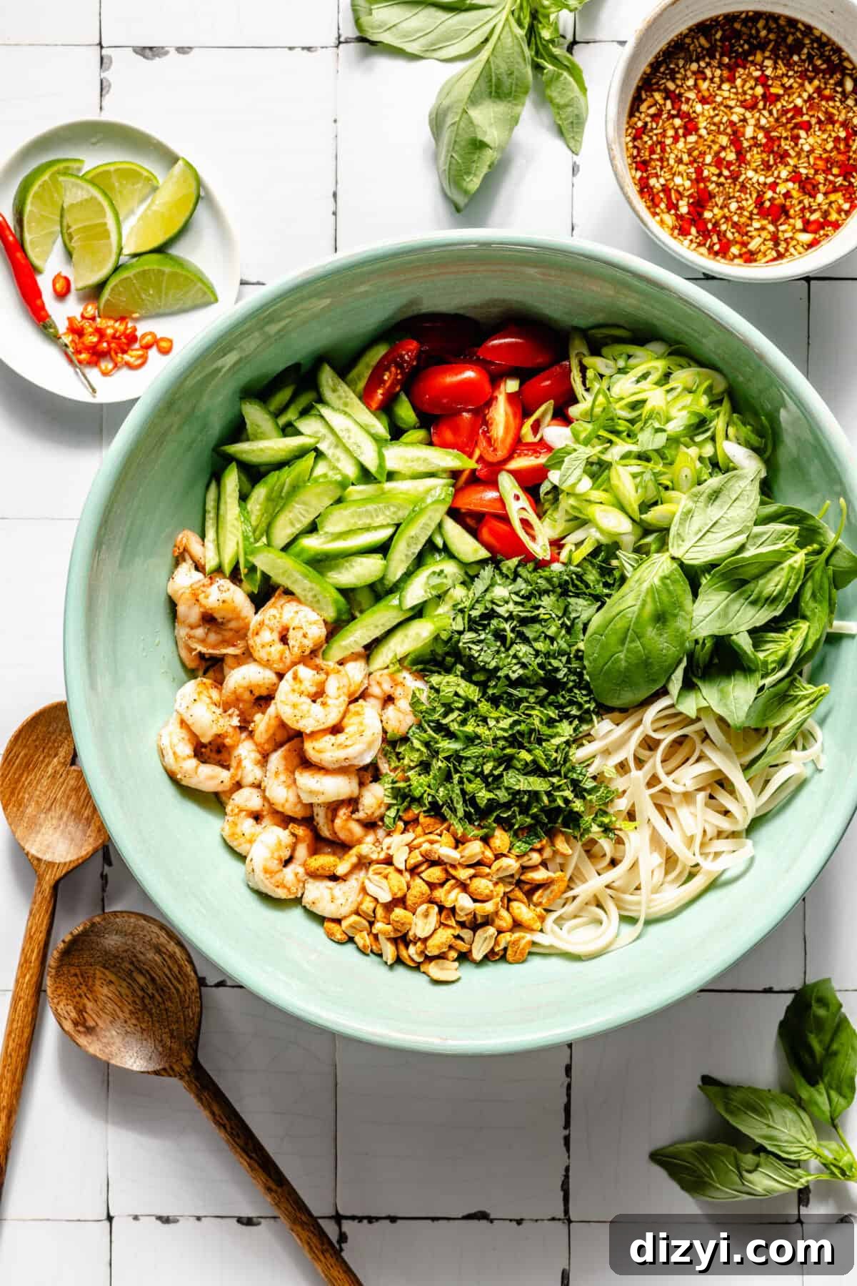 Sage green serving dish of Tangy Thai Shrimp Noodle Salad before it is mixed together. White bowl with dressing in the top right corner, basil leaves, and a dish of lime wedges and Thai chiles in the top left. Wooden tongs in the bottom left corner, as well as more basil leaves in the bottom right.
