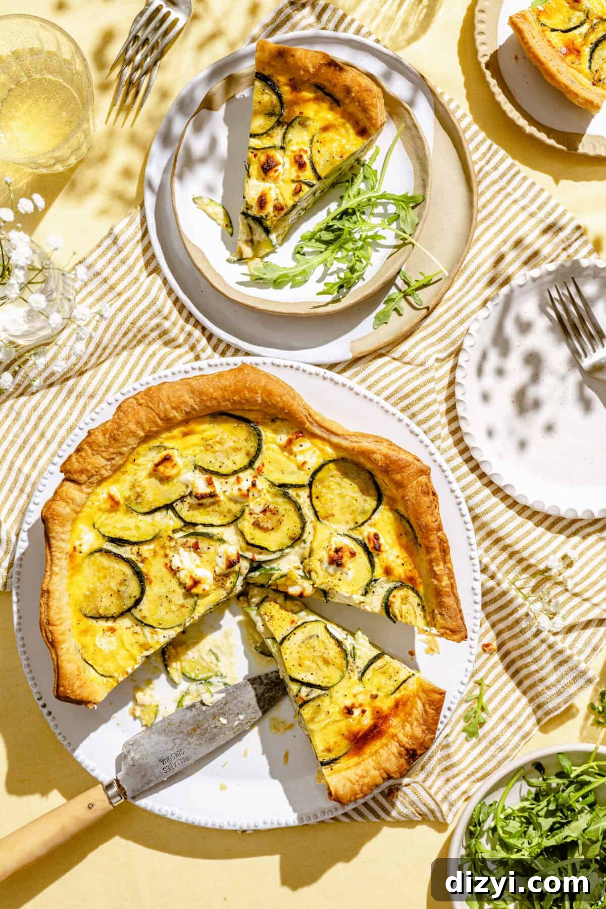 Zucchini and Goat Cheese Quiche with Flaky Puff Pastry 2 A serving dish with the finished quiche, with three slices cut out. One slice is on a white plate in the top right corner, one is featured at the top of the image along with some arugula, and the third slice remains on the serving dish. There is a empty white plate in the right of the image, along with a vase of baby's breath and a glass of water.