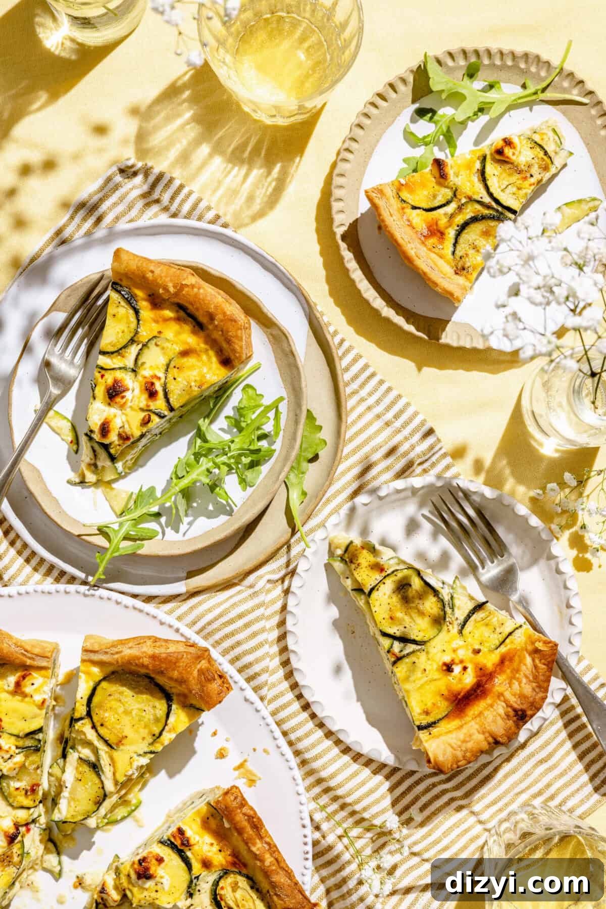 Zucchini and Goat Cheese Quiche with Flaky Puff Pastry 4 This image shows a tablescape with a yellow tablecloth serving Puff Pastry Zucchini and Goat Cheese Quiche. There are four small white plates, some forks, one of which has a slice of the quiche along some arugula. There is a small vase of baby's breath on the table, along with glasses of water.