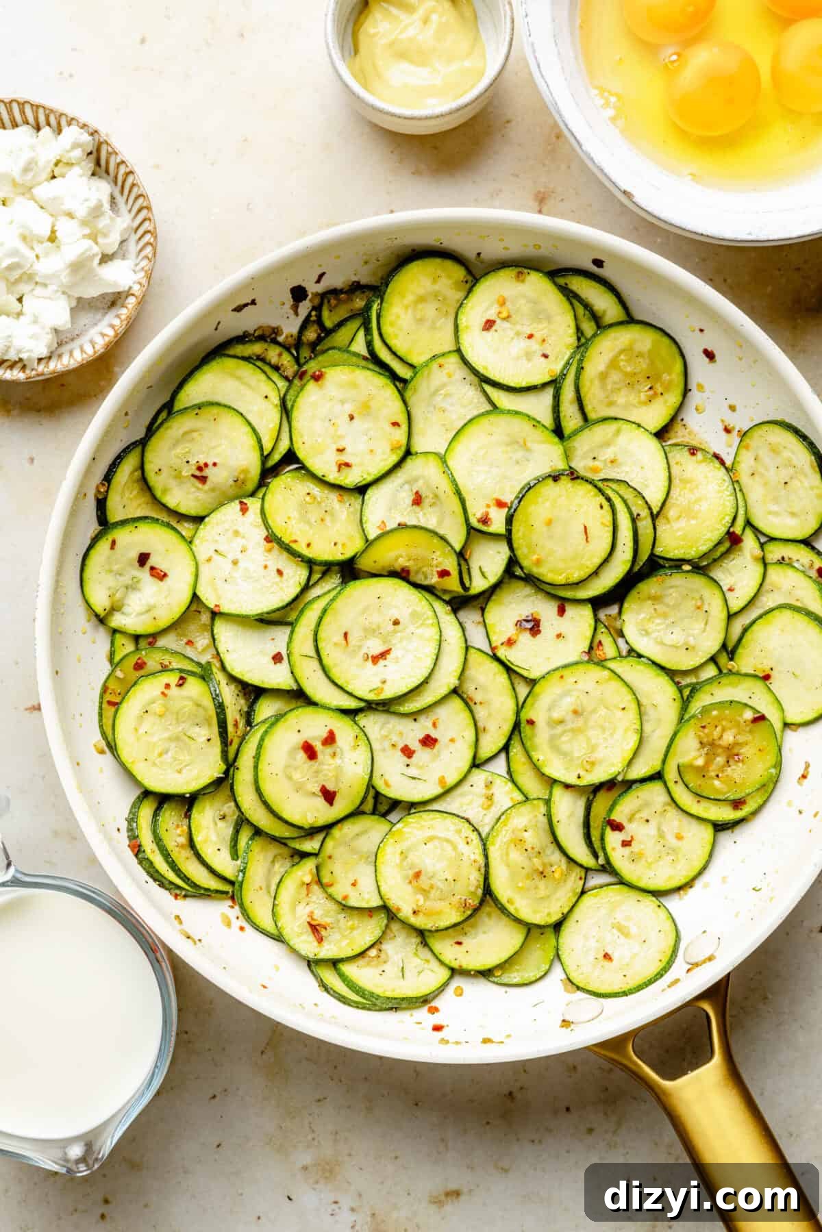 Zucchini and Goat Cheese Quiche with Flaky Puff Pastry 5 Thinly sliced zucchini in a white pan with a gold handle. In the top right corner is a bowl of freshly cracked eggs, next to a small white bowl of Dijon mustard. A brown and white bowl of crumbled goat cheese is in the top left corner, with a measuring cup of milk in the bottom left corner.