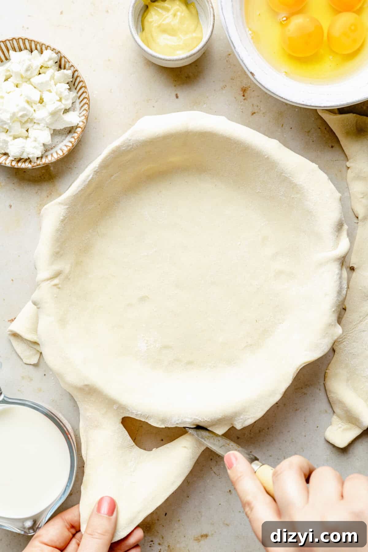 Zucchini and Goat Cheese Quiche with Flaky Puff Pastry 6 A layer of puff pastry is being trimmed to fit the dish. In the top right corner is a white bowl of freshly cracked eggs, next to a smaller white bowl of Dijon mustard. In the top left corner is a small brown and white bowl of crumbled goat cheese. In the bottom left is a measuring cup of milk.