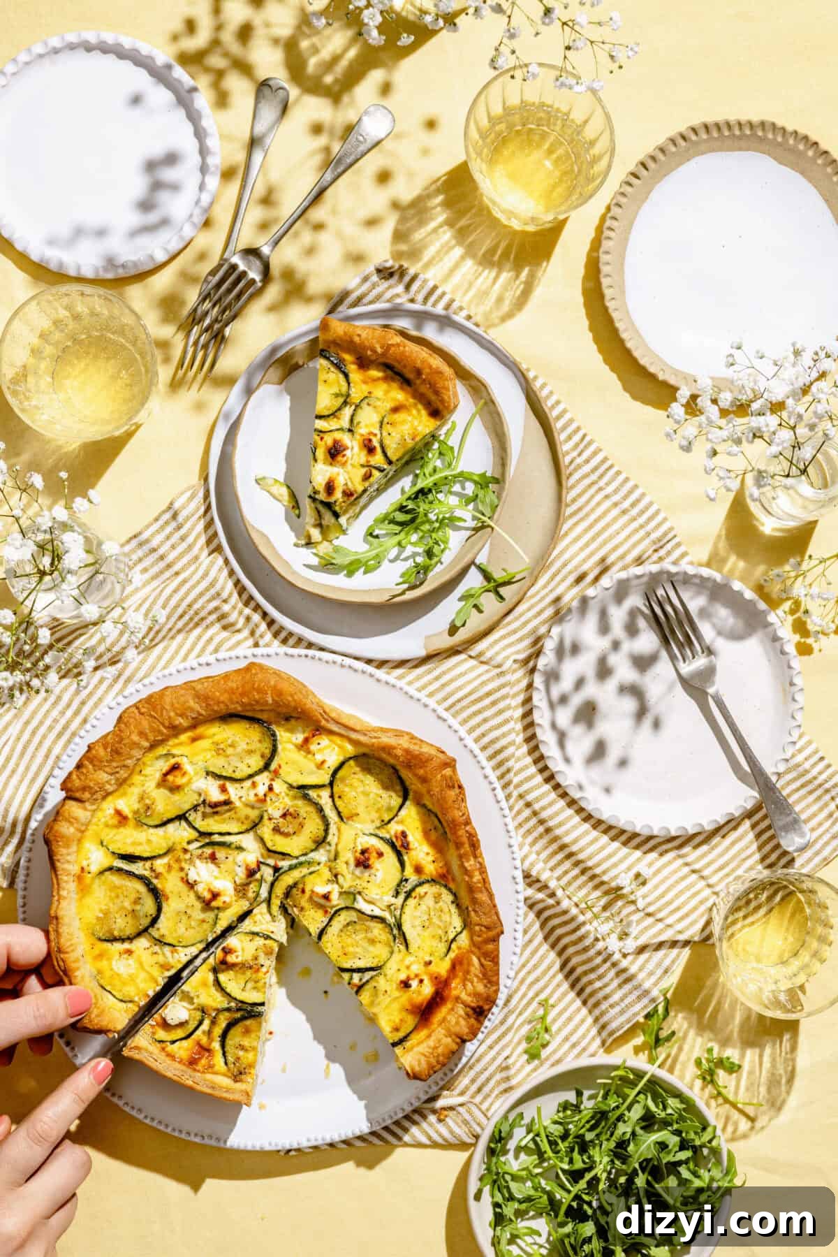 Zucchini and Goat Cheese Quiche with Flaky Puff Pastry 7 This image shows a tablescape with a yellow tablecloth serving Puff Pastry Zucchini and Goat Cheese Quiche. There are four small white plates, some forks, one of which has a slice of the quiche along some arugula. There are small vases of baby's breath on the table, along with glasses of water. A small white bowl of arugula is in the bottom right corner.