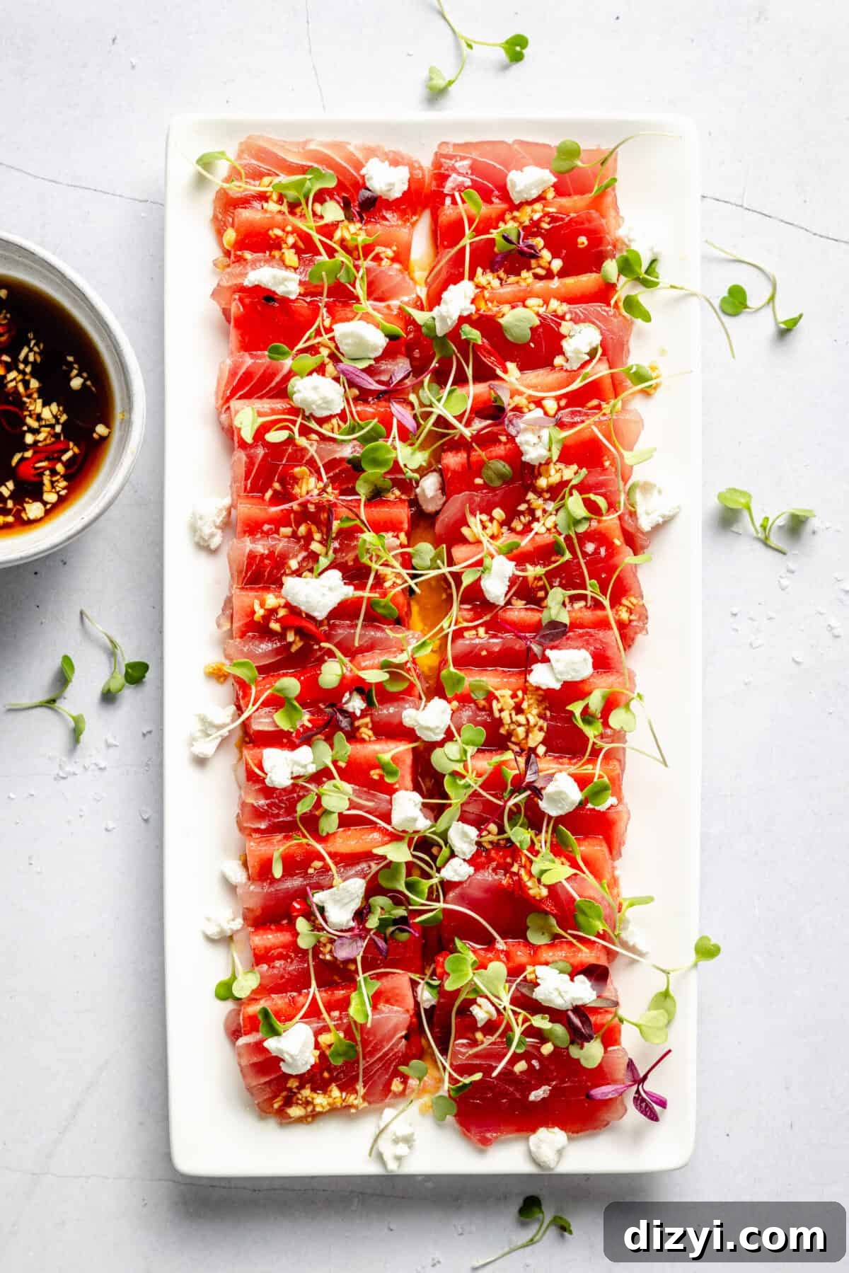 Watermelon and Tuna Sashimi Recipe 2 Top-down shot of Watermelon and Tuna Sashimi on a white rectangular platter, layered with ahi tuna, watermelon, microgreens, and crumbled goat cheese, with a bowl of ponzu sauce nearby.