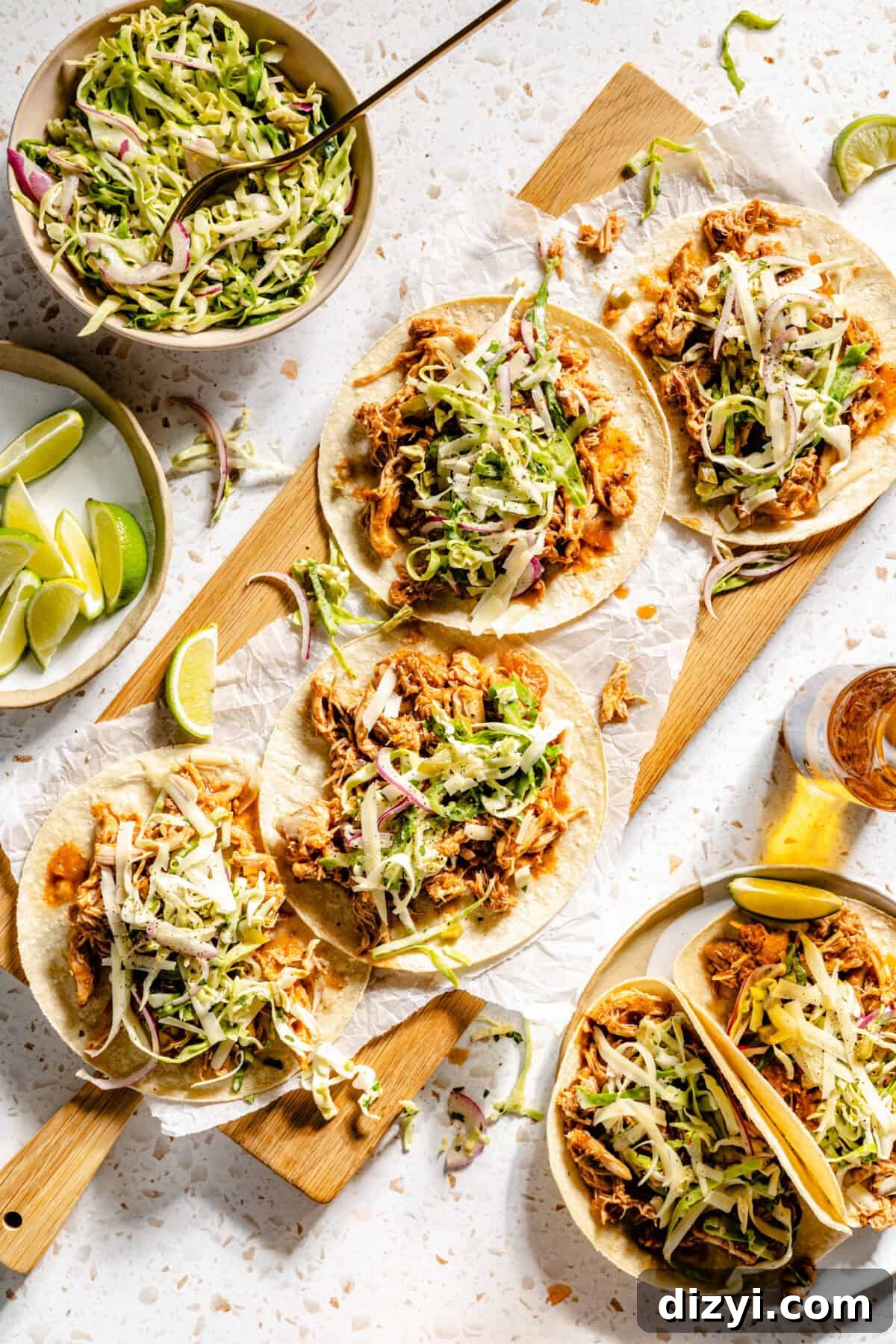 Slow Cooker BBQ Chicken Tacos with Shredded Chicken 2 The Slow Cooker BBQ Chicken Tacos assembled on a serving board, a bowl of slaw on the side, a dish with lime wedges, and a plate with two additional tacos.