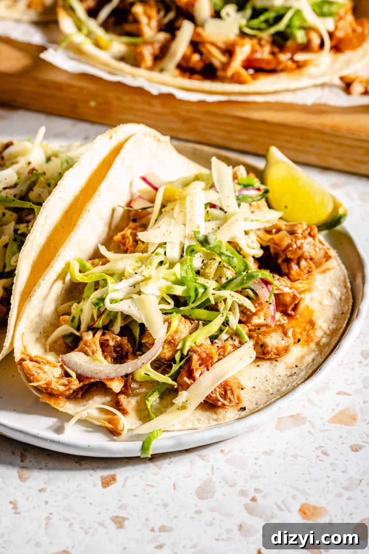 Slow Cooker BBQ Chicken Tacos with Shredded Chicken 5 A close-up shot of two finished Slow Cooker BBQ Chicken Tacos served on a white plate with a lime wedge. A serving board with more tacos is in the background.