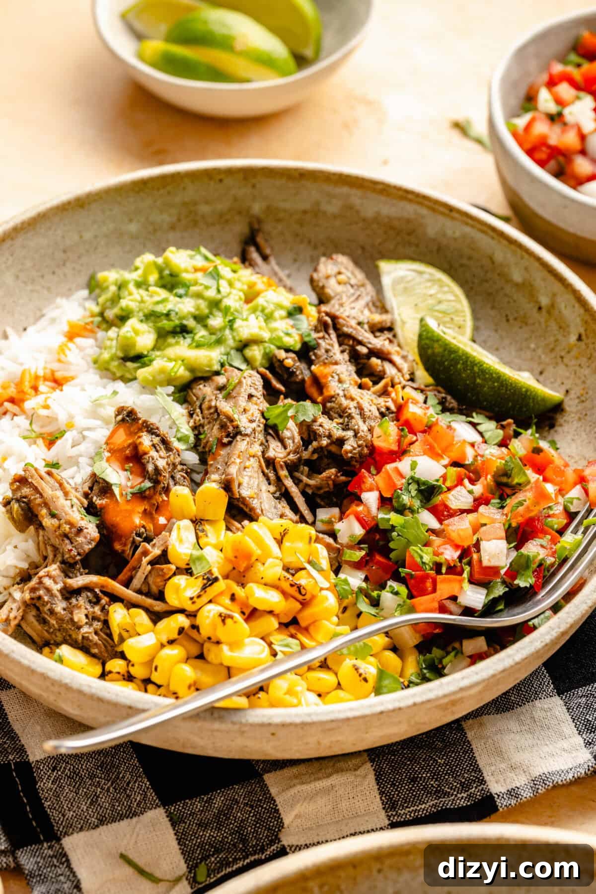 A finished Shredded Beef Chipotle Burrito Bowl served in a tan-colored dish with a fork on top of a black and white checkered napkin. A small dish of lime wedges and a small dish of pico de gallo are also on the table.