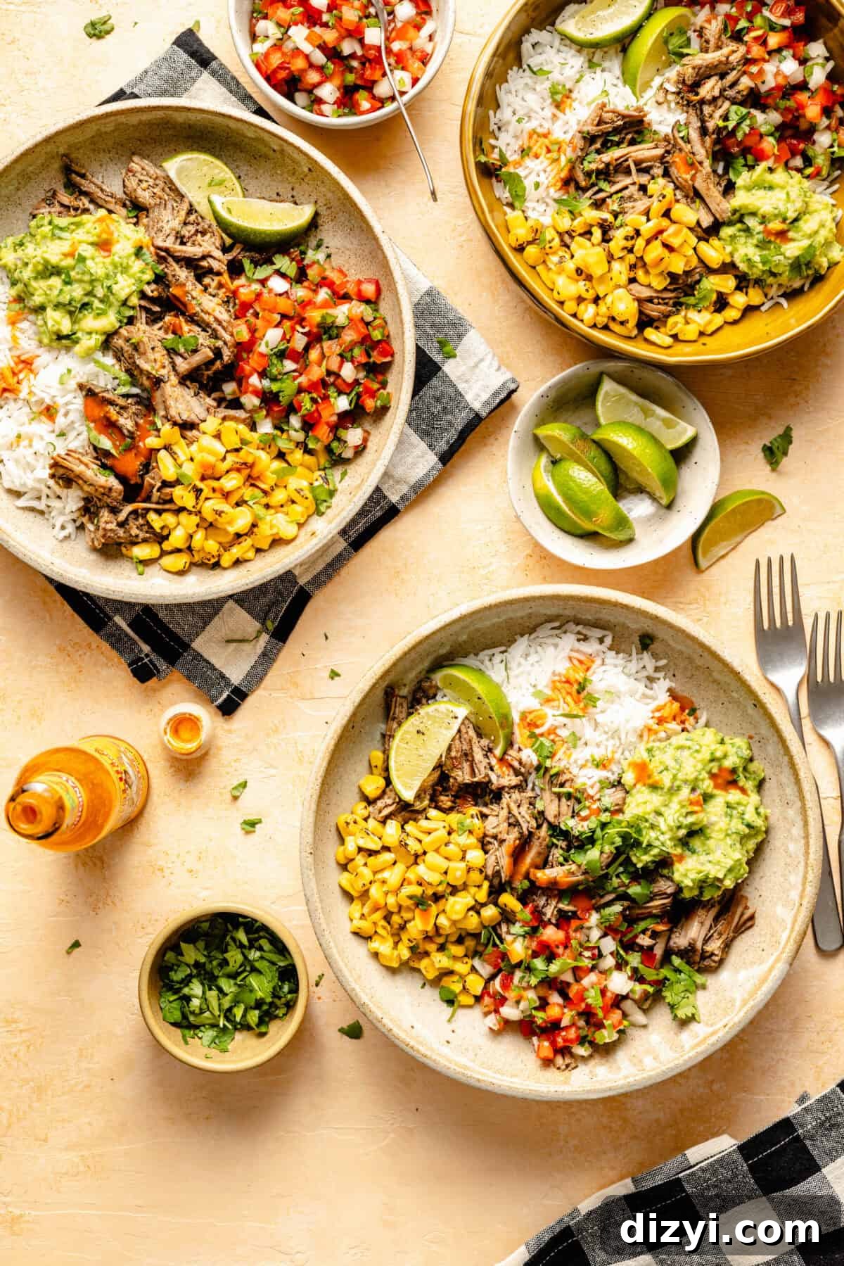 Three completed Shredded Beef Chipotle Burrito Bowls, served in cream-colored dishes. A small dish of cilantro, an open bottle of hot sauce, a bowl of limes, and a dish of pico de gallo are also on the table.
