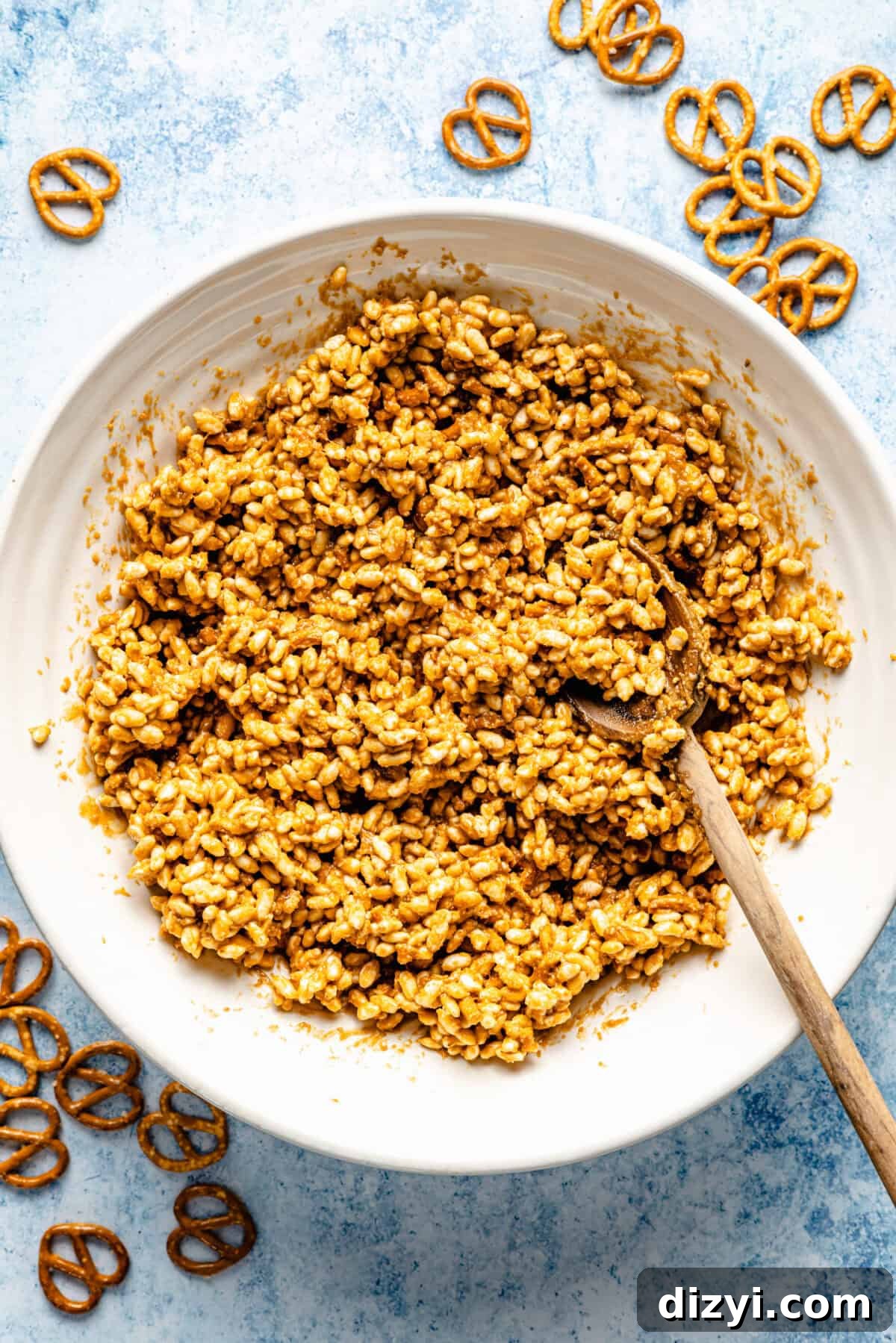 The peanut butter mixture with the cereal and pretzels added in a white bowl with a wooden spoon.