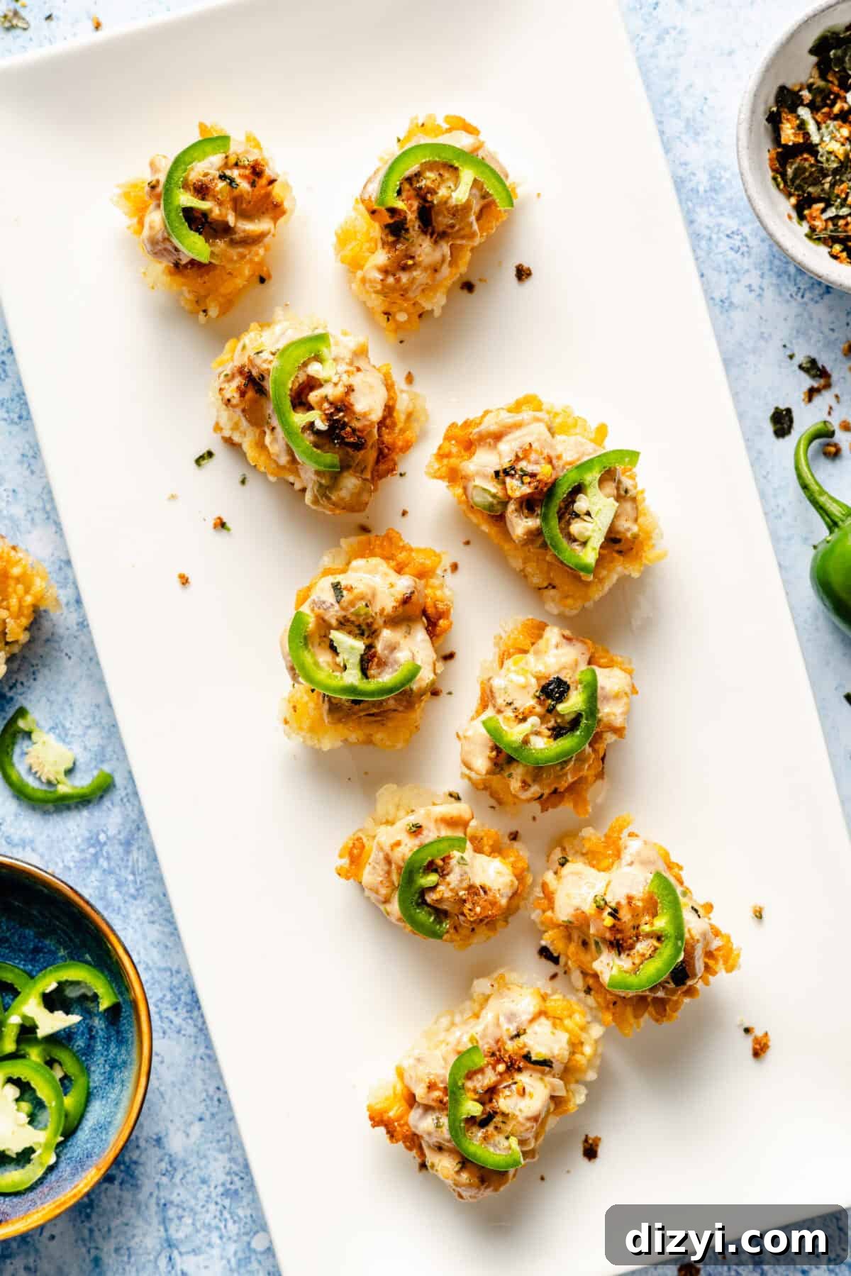 A white rectangular serving dish with the finished Spicy Tuna Crispy Rice. The dish sits on a blue countertop, with a small dish of spices, and sliced jalapenos also on the counter.