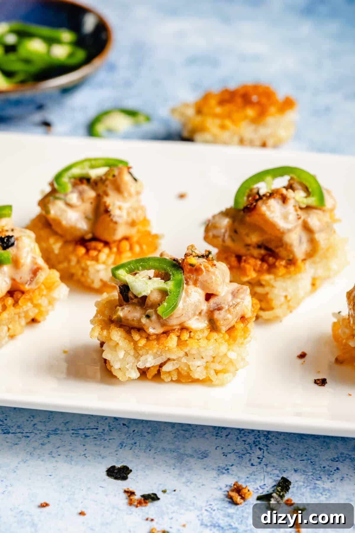 A close-up shot white rectangular serving dish with the finished Spicy Tuna Crispy Rice. The dish sits on a blue countertop, with a small dish of spices, and sliced jalapenos also on the counter.