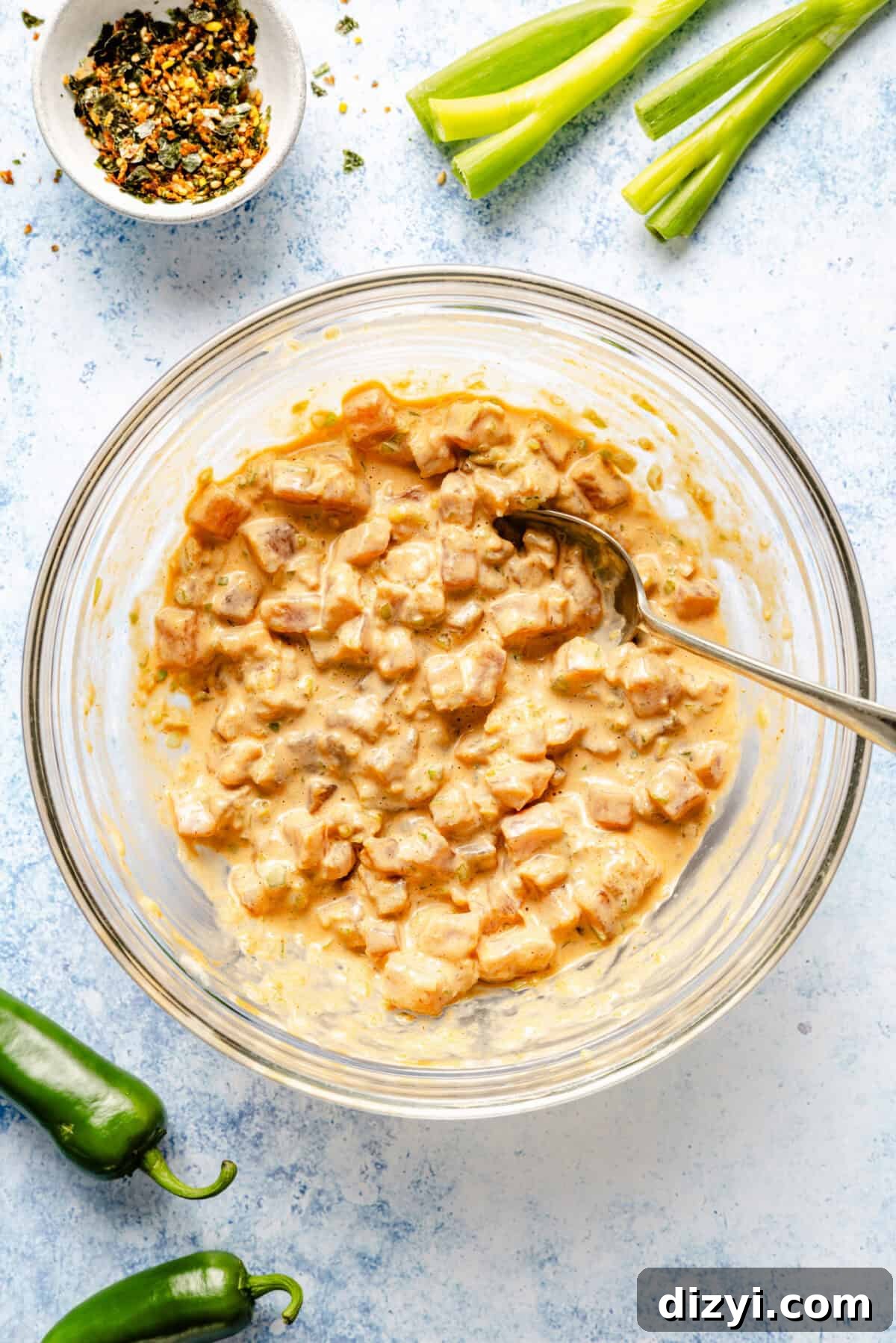 A clear glass bowl of the spicy tuna and sauce mixture sitting on a blue countertop. A white dish of spices, green onions, and jalapenos also on the counter.