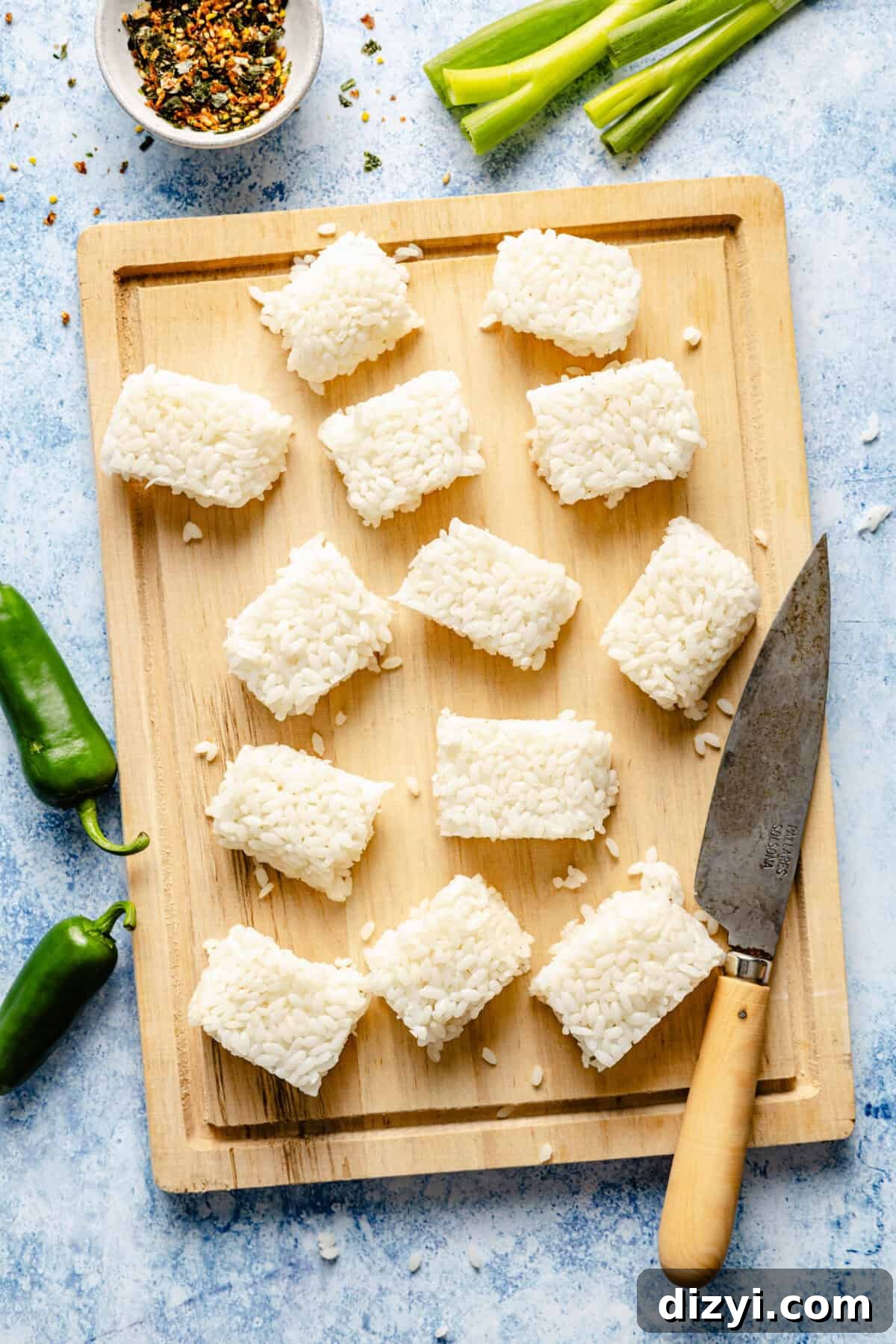 A wooden cutting board with rice cut into rectangles. A knife sits on the edge of the cutting board. Green onions, jalapenos, and a white dish of spices are also on the countertop.