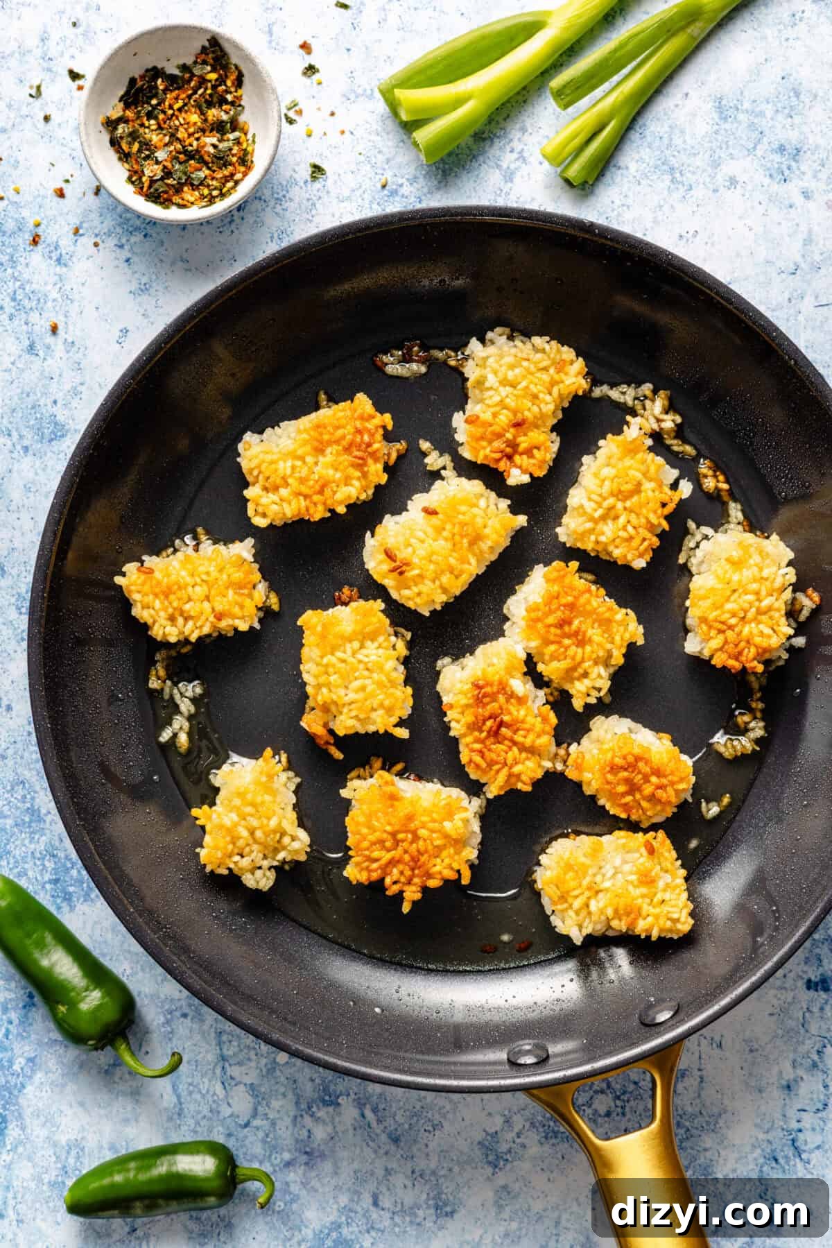 A black pan with a gold handle cooking the crispy rice. The pan is on a blue countertop, and a small dish of spices, green onions, and jalapenos are also on the counter.