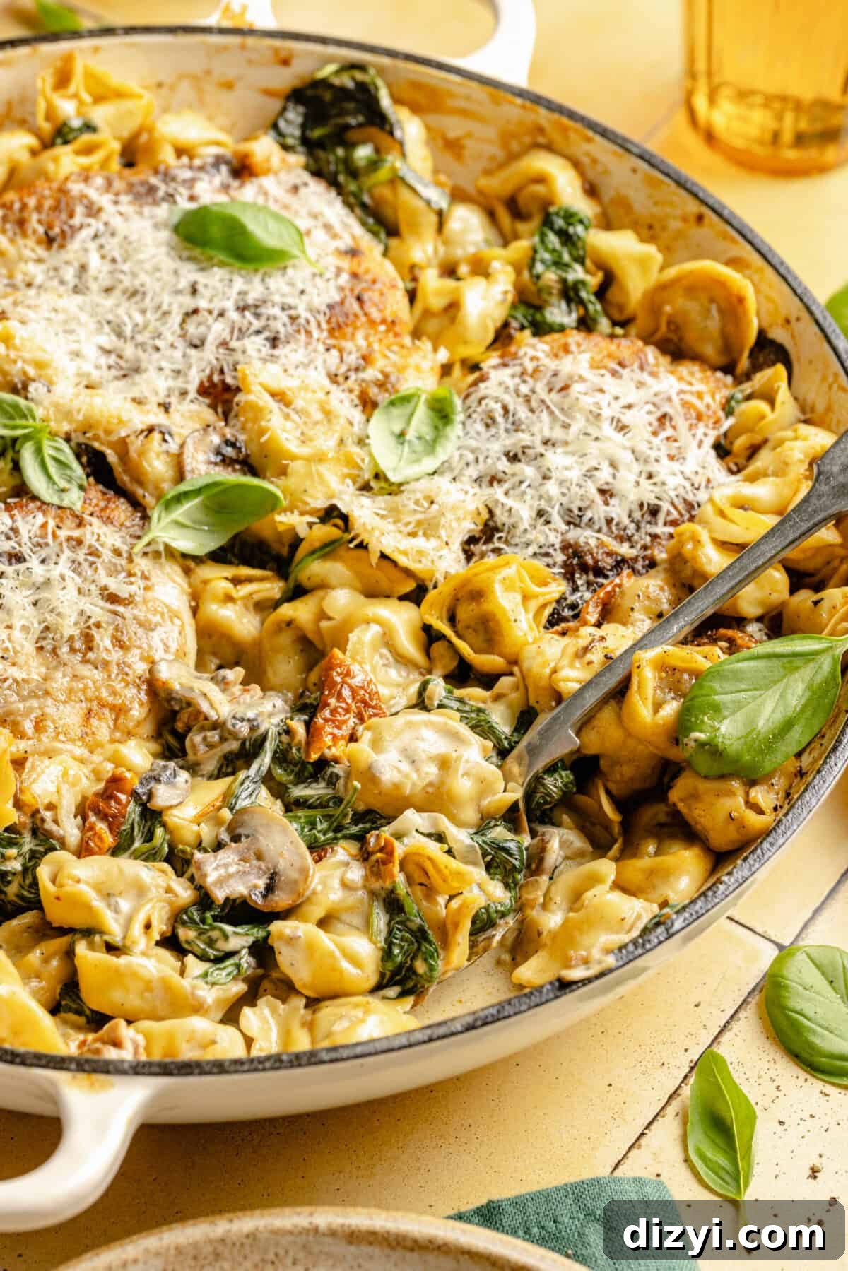 One-Pan Creamy Chicken and Tortellini Bake 2 A close-up shot of a pot of Creamy Spinach Chicken and Tortellini Bake on a yellow countertop. There is a serving spoon in the pot, a dark green napkin in the bottom right corner.