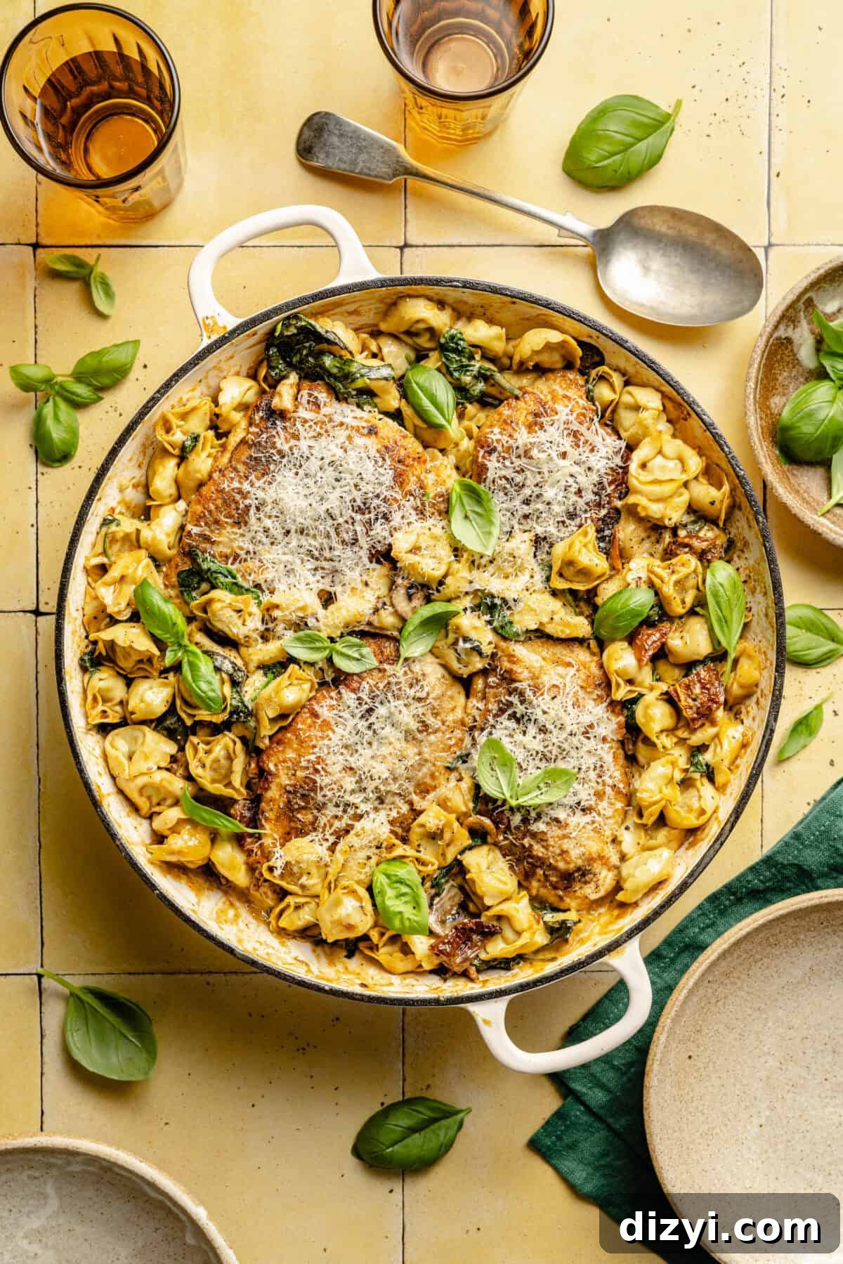 One-Pan Creamy Chicken and Tortellini Bake 5 A pot of Creamy Spinach Chicken and Tortellini Bake on a yellow countertop. There is a serving spoon at the top of the image, a dark green napkin in the bottom right corner, as well as tan dishes. Two amber-colored glasses in the top of the image, and basil leaves spread throughout the image.
