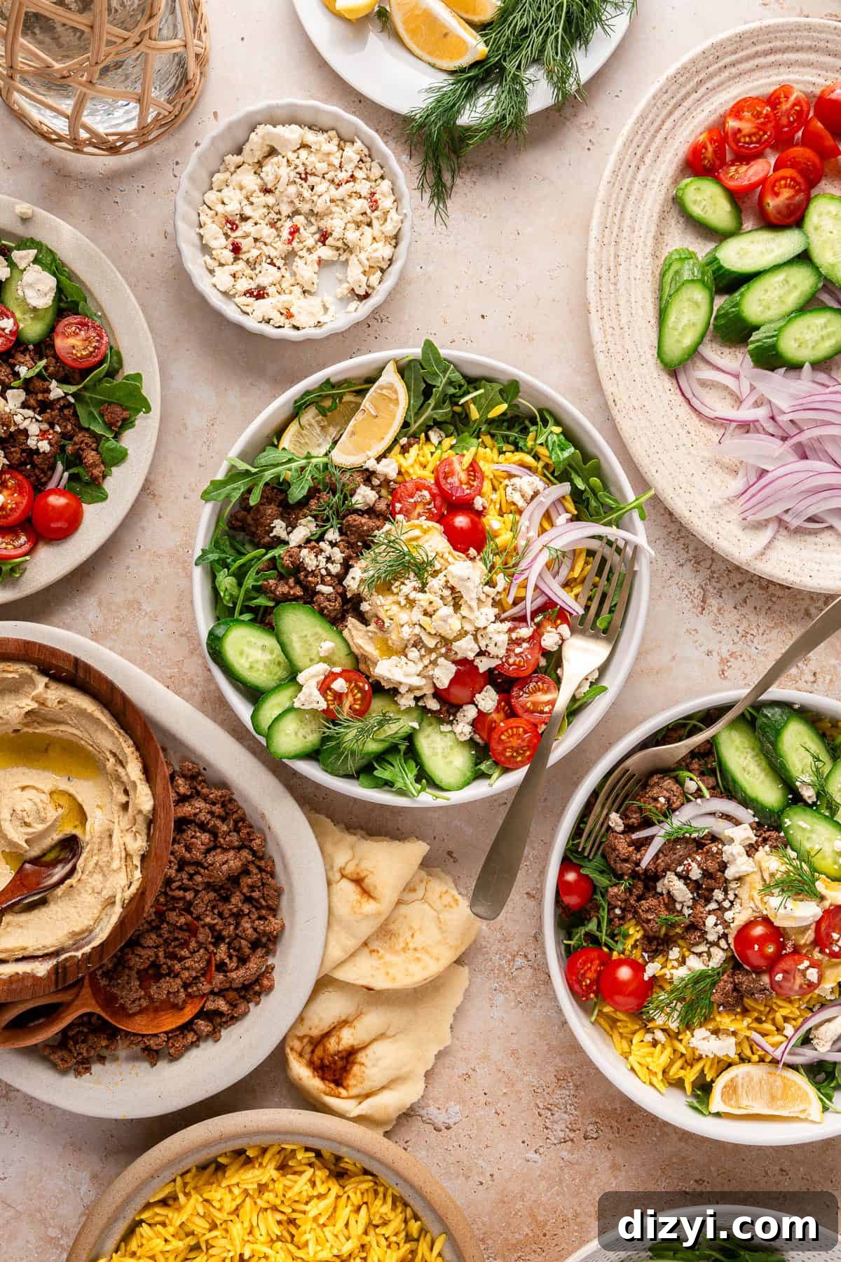 Assembled Ground Lamb Hummus Bowls, surrounded by dishes of other toppings, dill, lemon slices, rice, hummus, and ground lamb.