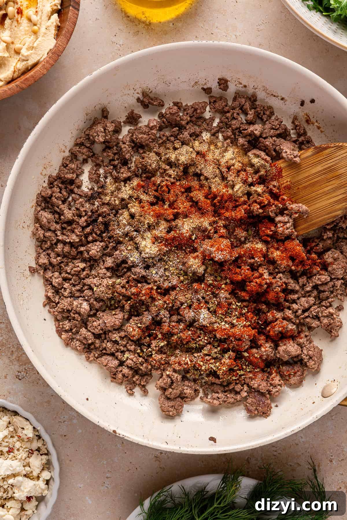 The ground lamb and seasonings being cooked in a white pan with a wooden spoon.