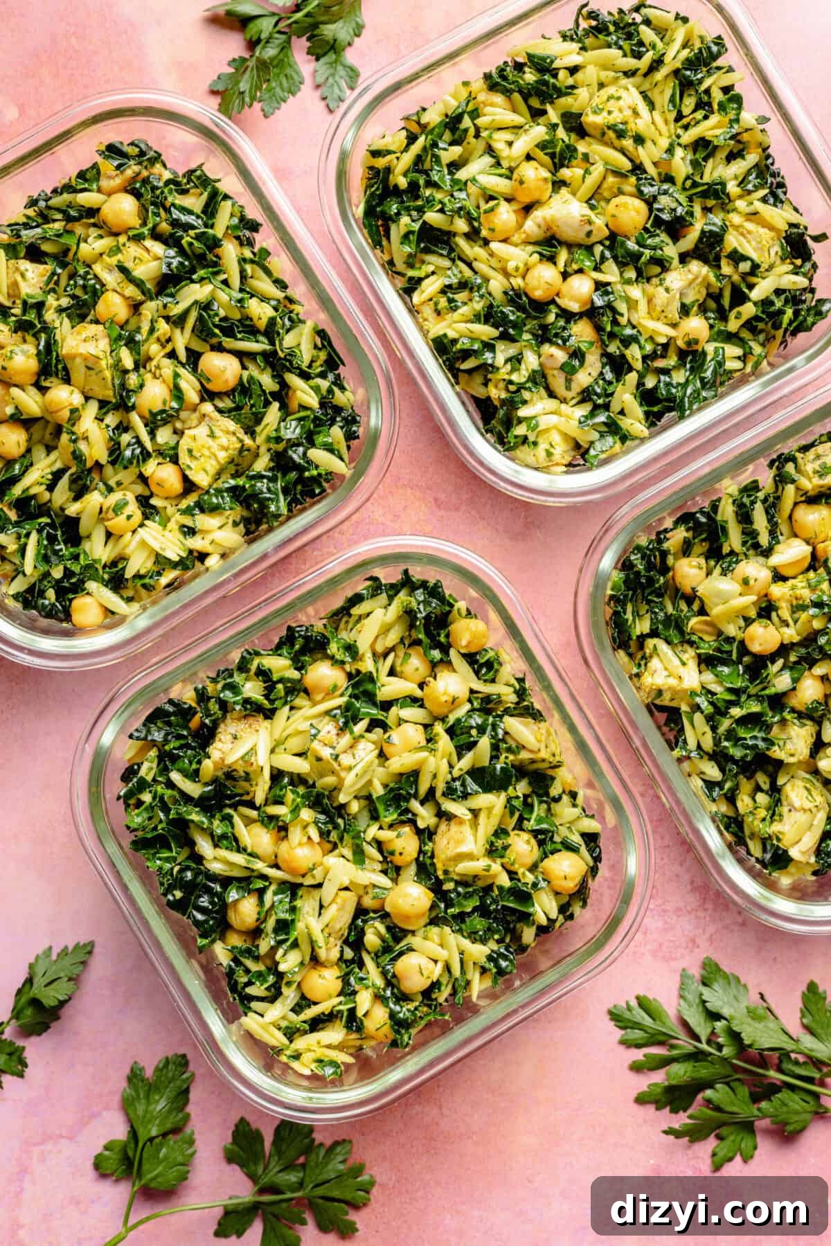 Four meal prep containers with Lemon Chicken, Orzo, and Chickpea Salad on a pink countertop.
