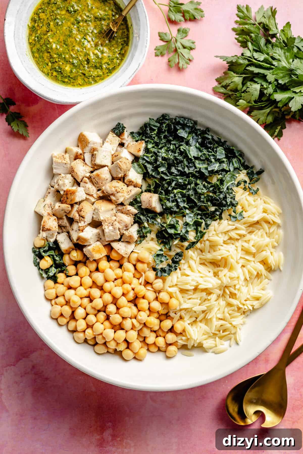 The finished ingredients for Four meal prep containers with Lemon Chicken, Orzo, and Chickpea Salad on a pink countertop a white bowl, waiting to be tossed with the dressing. The dressing sits in a small white dish to the side. The dishes are on a pink countertop.