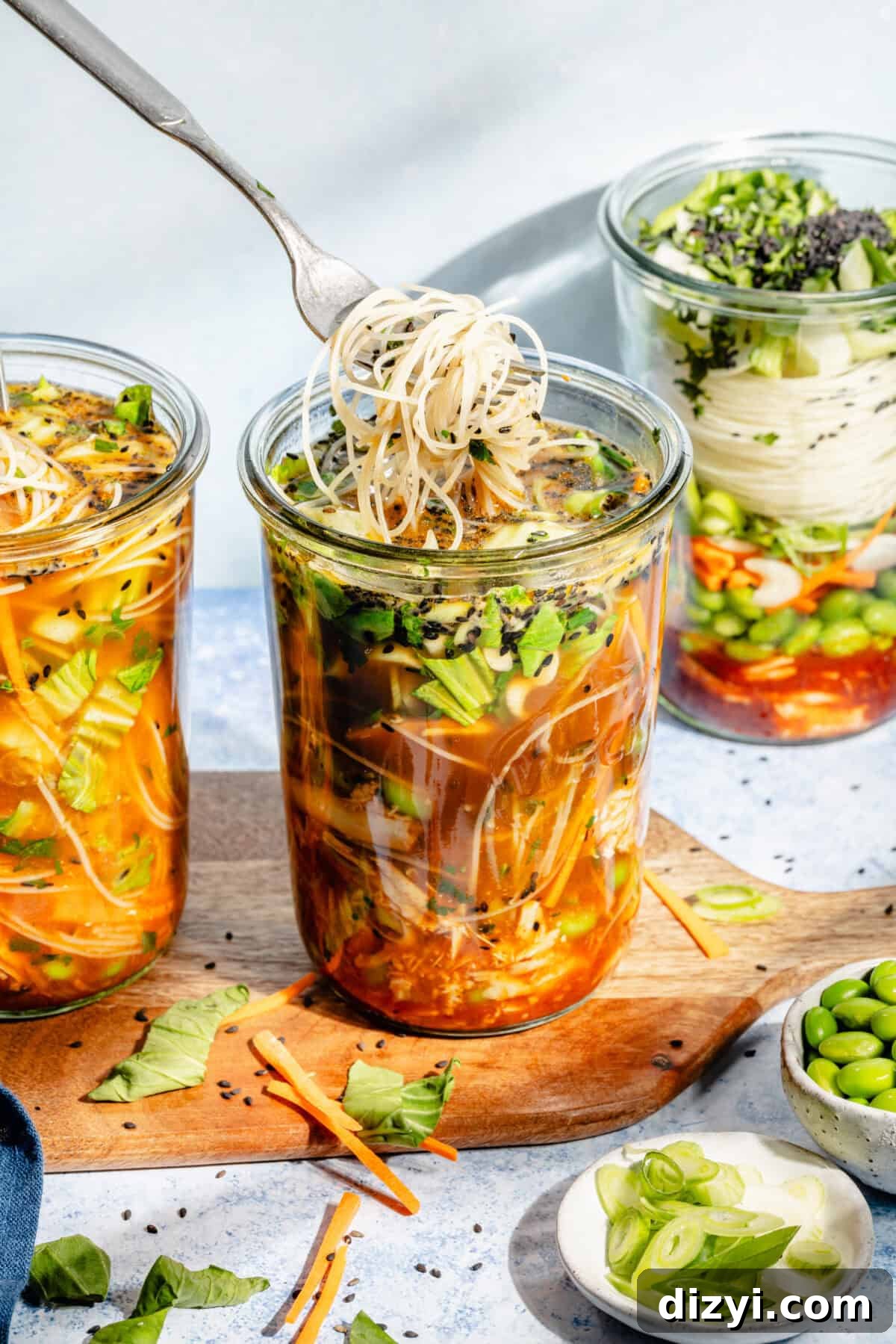 Three completed Mason Jar Instant Gochujang Noodles on a wooden serving board. Various other ingredients surround them in small dishes. A fork is visible twirling some noodles.