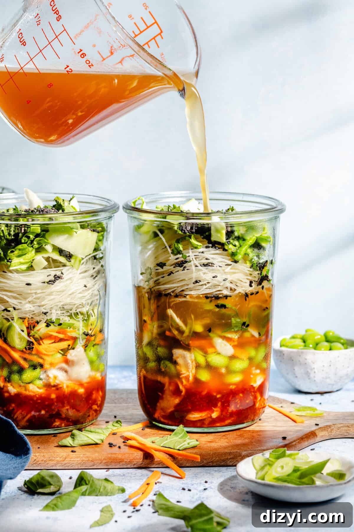 Three completed Mason Jar Instant Gochujang Noodles on a wooden serving board. Various other ingredients surround them in small dishes. The dishes are on a blue counter. Broth is being poured into the mason jars from a clear glass measuring cup.