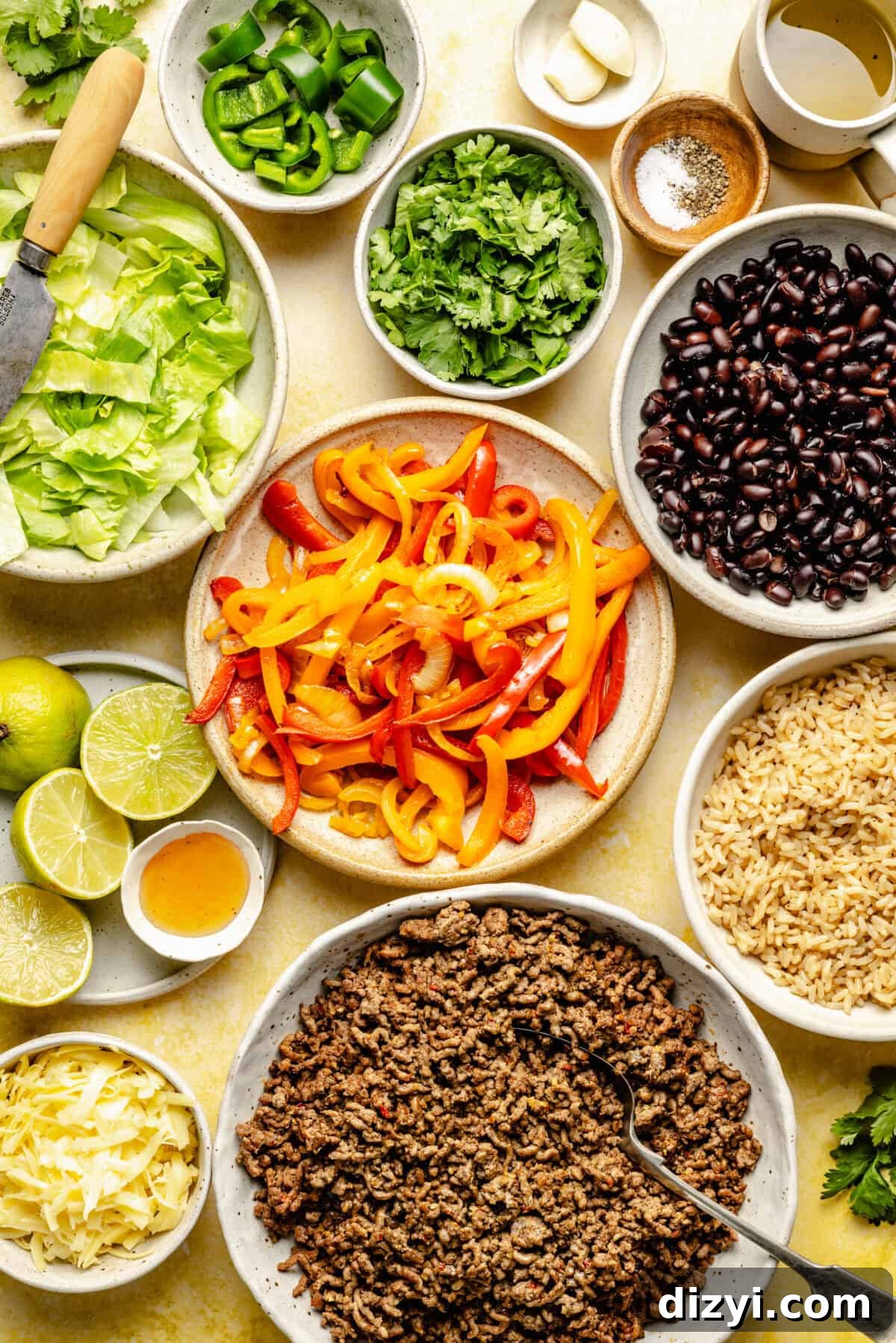 Meal-Prep Taco Bowls with Jalapeno Dressing 4 The various ingredients needed to make Make Ahead Taco Bowls spread on a table - a bowl of red and yellow bell peppers, ground beef, brown rice, black beans, cilantro, jalapenos, cheese, lettuce, and limes.
