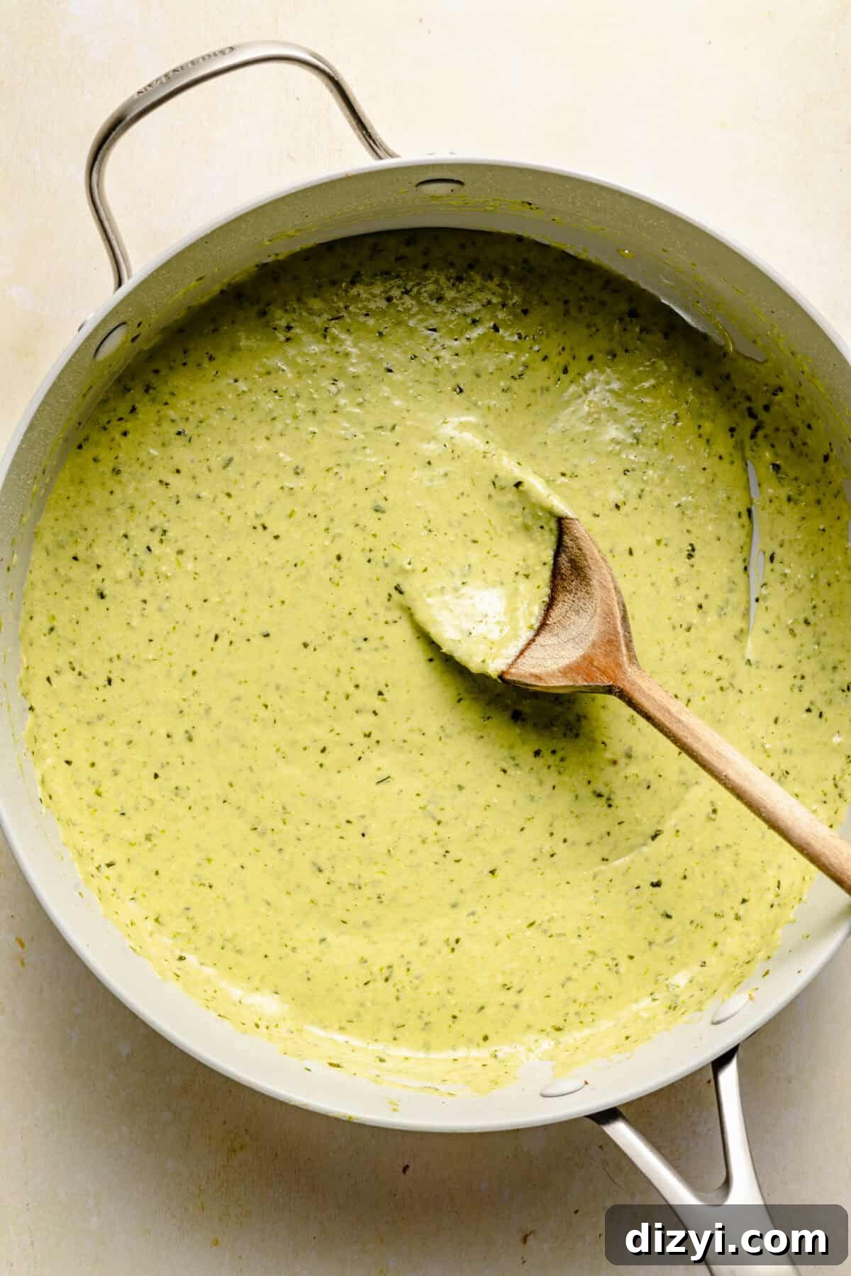 The creamy pesto sauce in a skillet with a wooden spoon on a white countertop.